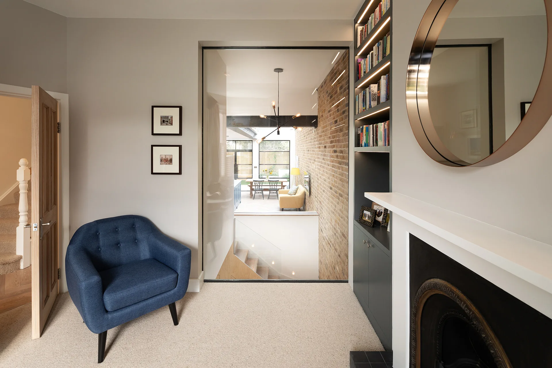 A blue armchair sits on a carpeted floor next to a fireplace and a large interior glass wall looking into an adjacent dining area.