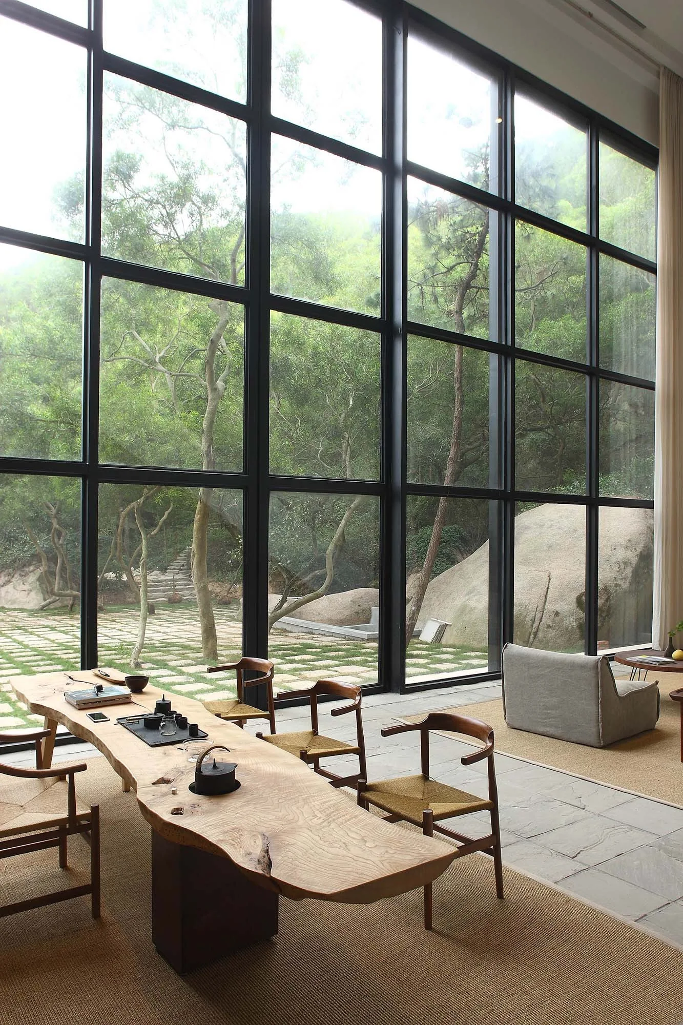 A minimalist tea room with a long live-edge wood table, woven-seat chairs, and massive black grid windows overlooking a garden.