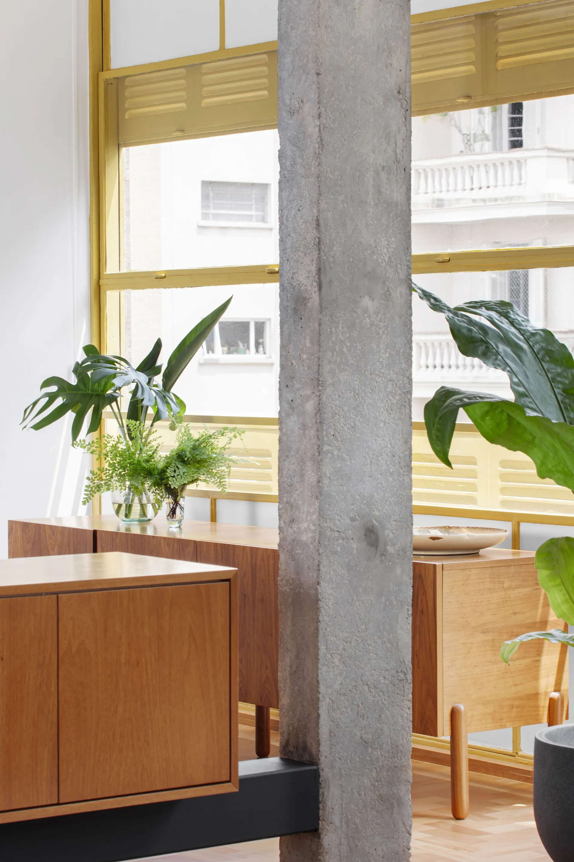 A bright room featuring mid-century wooden sideboards, houseplants in glass vases, a structural concrete pillar, and yellow window frames.