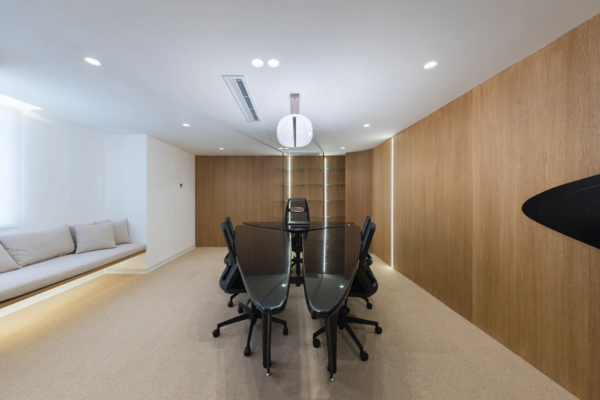 A modern meeting room features a black glossy sculptural conference table surrounded by mesh office chairs, a built-in bench, and wood paneled walls.