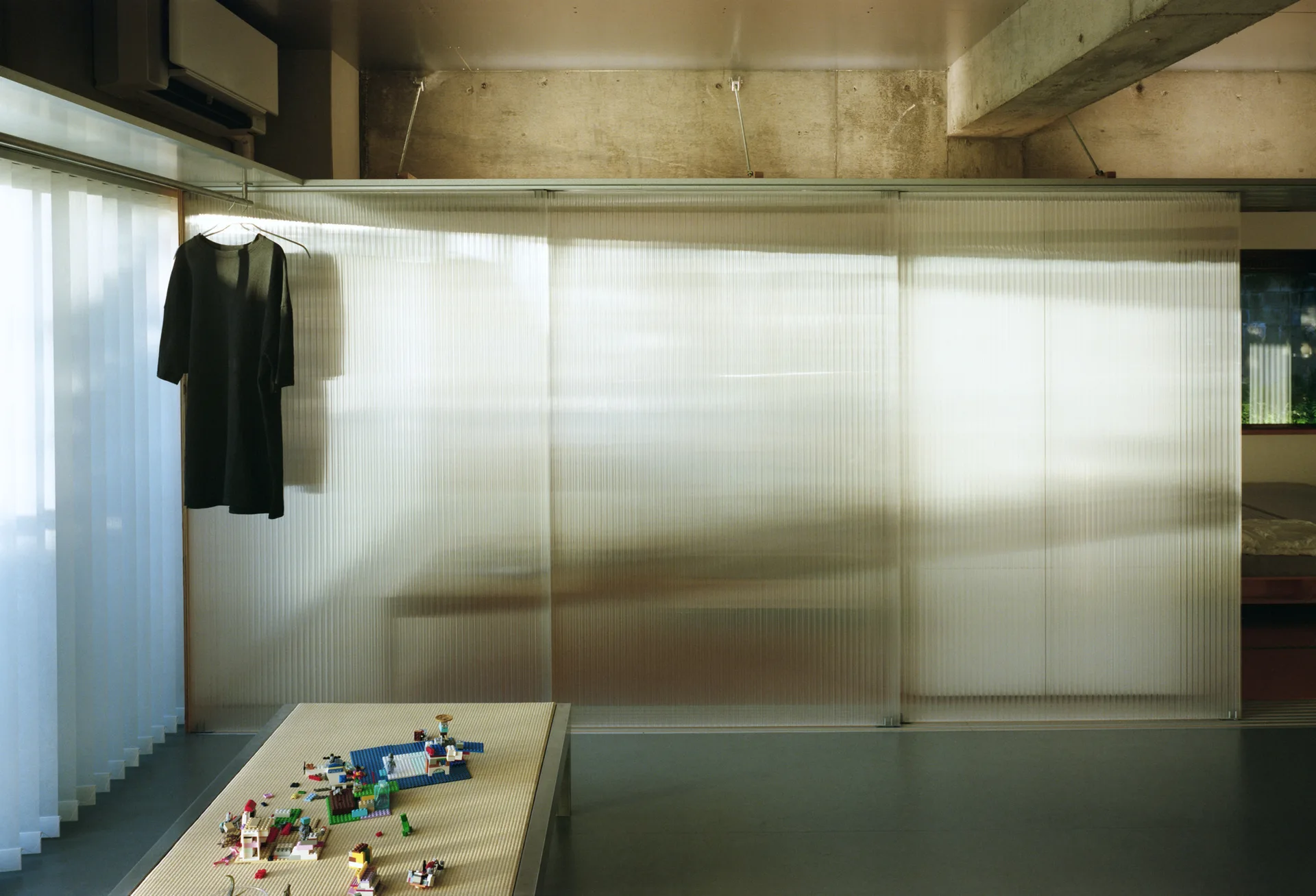 Translucent fluted sliding partition walls divide a minimalist room with a low tatami table and exposed concrete ceiling.