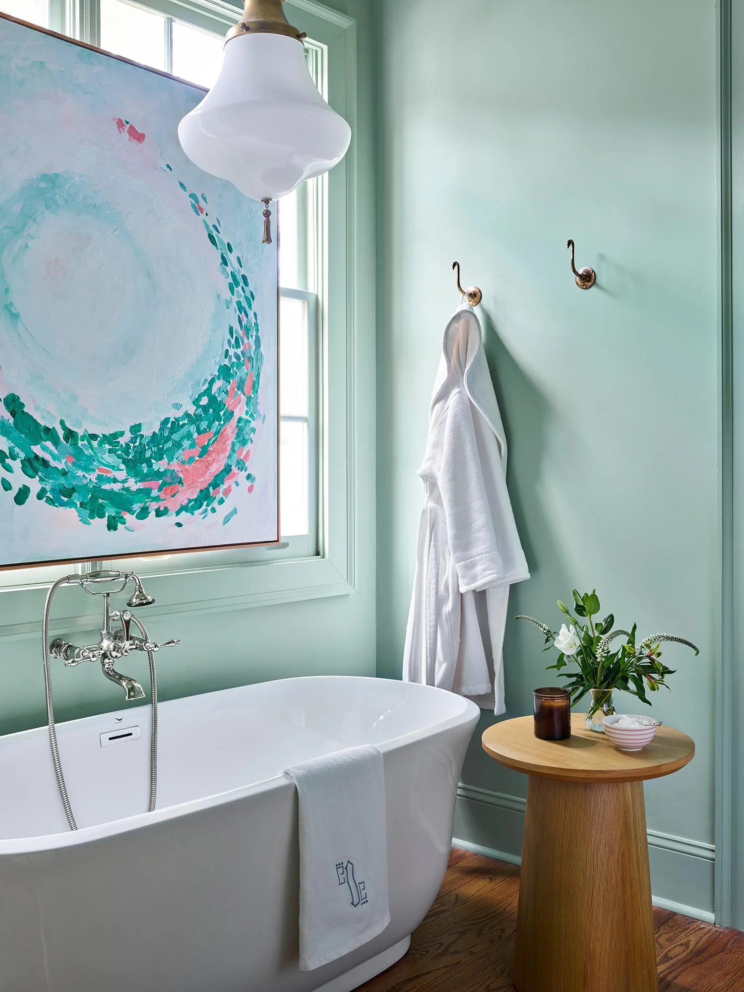 A bathroom with mint green walls features a white freestanding bathtub under a window, a wooden side table, and a white robe hanging on brass hooks.