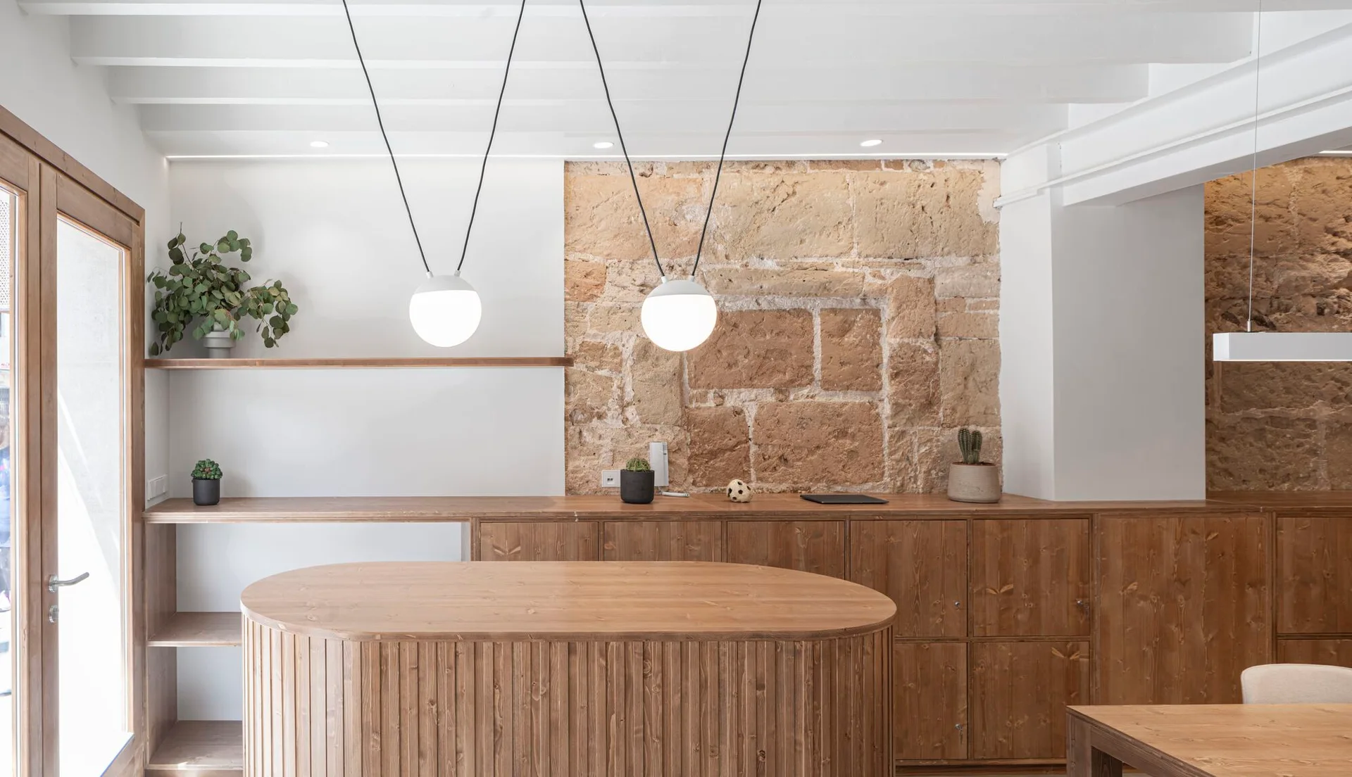 A minimalist wooden kitchen features a central oval island, matching low cabinets, spherical pendant lights, and an exposed rough stone accent wall.