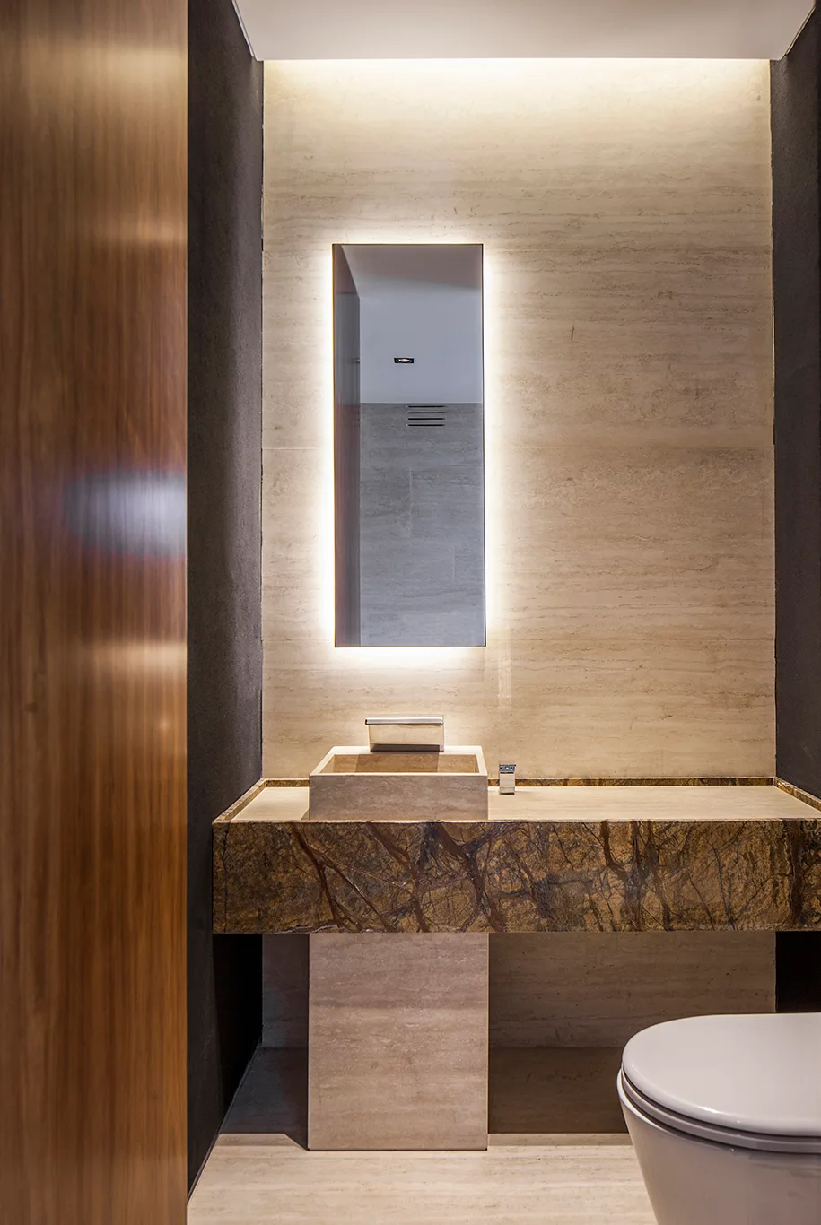 A modern powder room featuring a stone vanity with a rectangular vessel sink, a tall backlit mirror, and beige travertine walls.