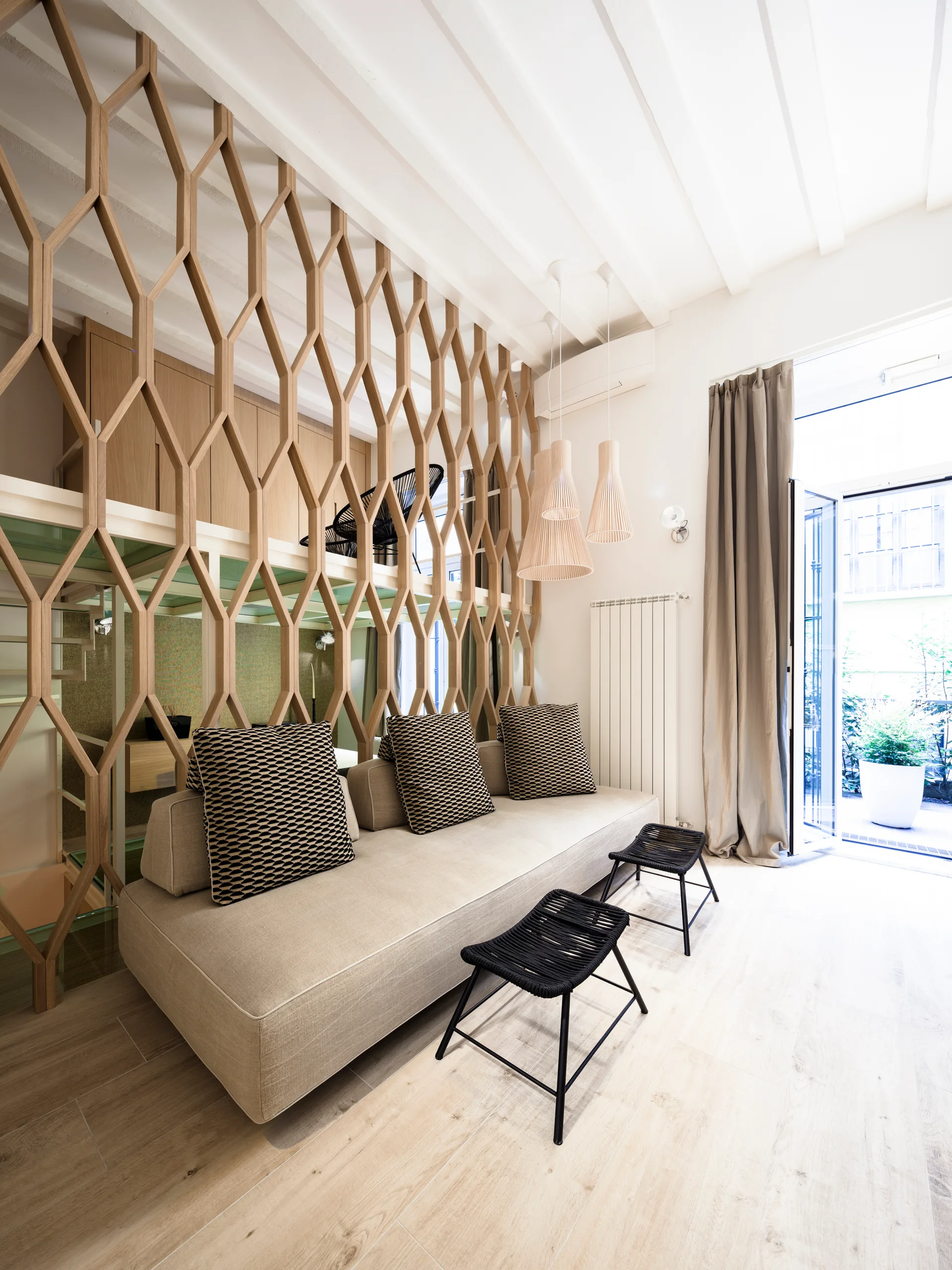 A light-toned living room features a beige sofa with geometric pillows, black woven stools, and a large honeycomb wooden screen partition.