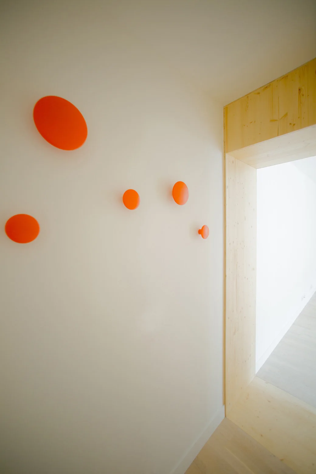Five vibrant orange round wall hooks of varying sizes are mounted on a white wall beside a light wood doorframe.
