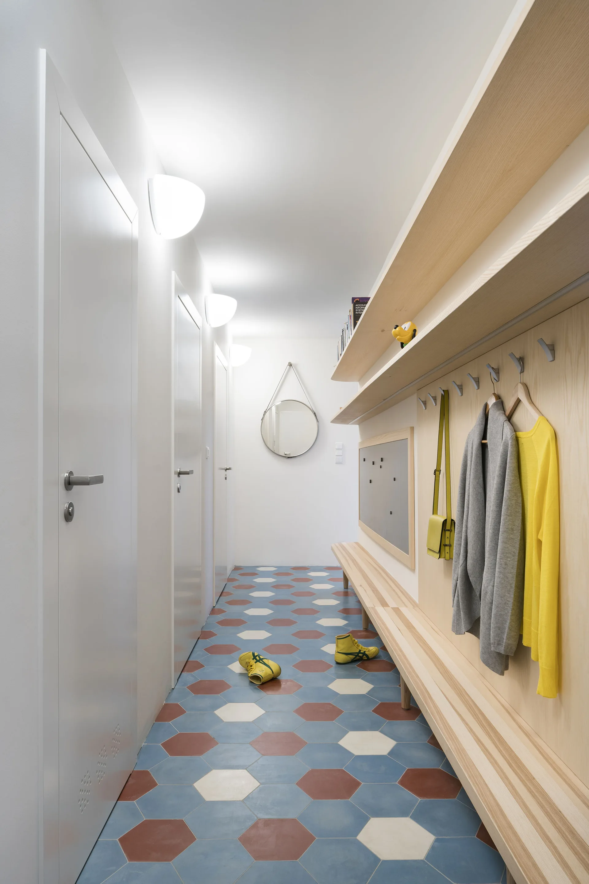 A modern hallway featuring blue and terra cotta hexagonal floor tiles, a long light wood built-in bench, and white walls with sconces.