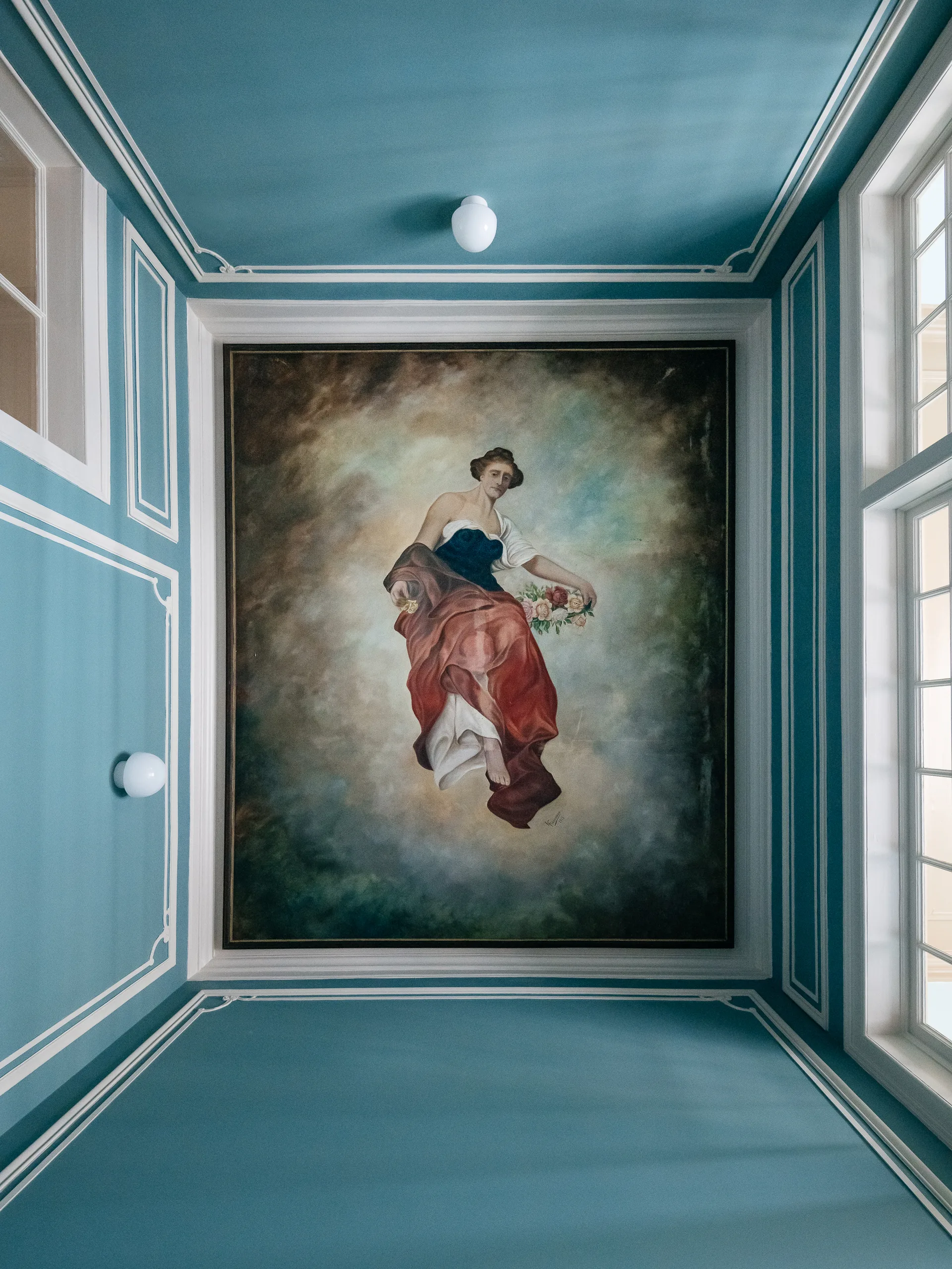 A low-angle view of a teal-blue ceiling and walls decorated with delicate white panel moulding and a large classical painting of a woman in the center.
