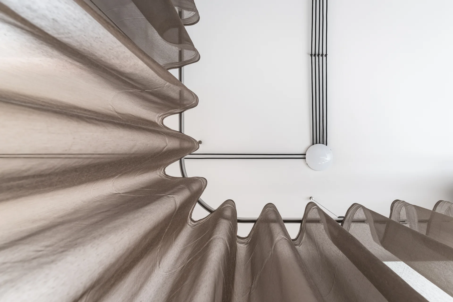 A low-angle view of sheer brown curtains hanging from a curved ceiling track alongside black electrical conduits on a white ceiling.
