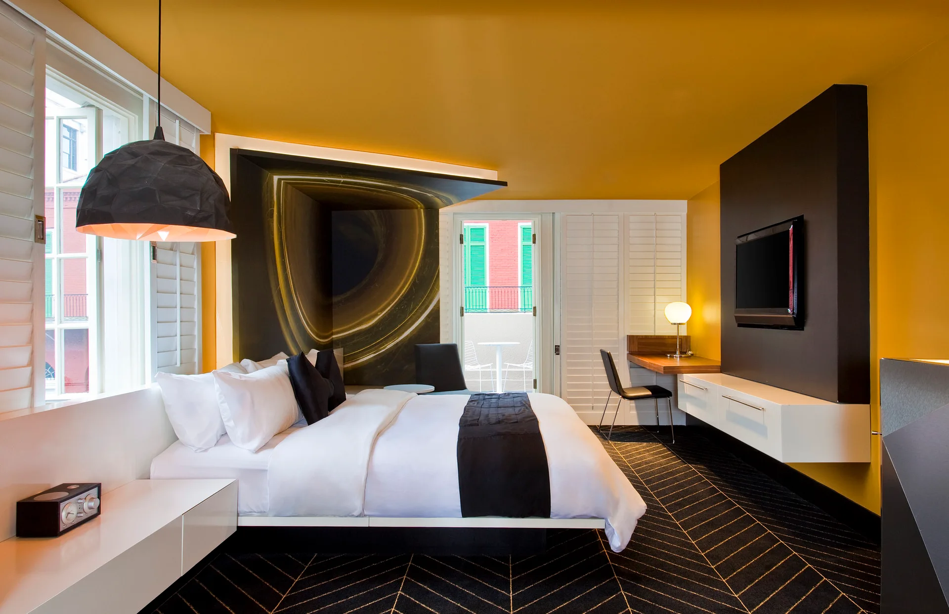 A modern bedroom features a mustard yellow ceiling, white bedding, white plantation shutters, and a floating storage unit under a wall-mounted TV.