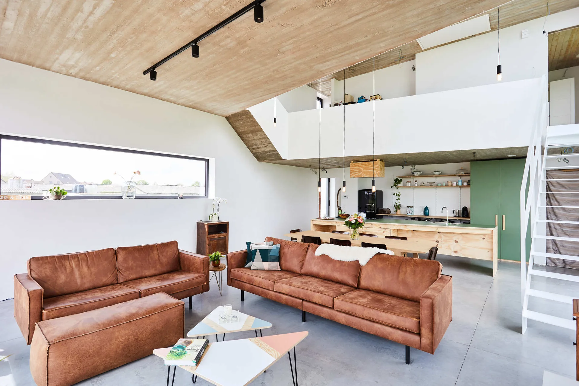 An open plan loft living room features two brown leather sofas, a textured concrete ceiling, and a kitchen with green cabinets in the background.