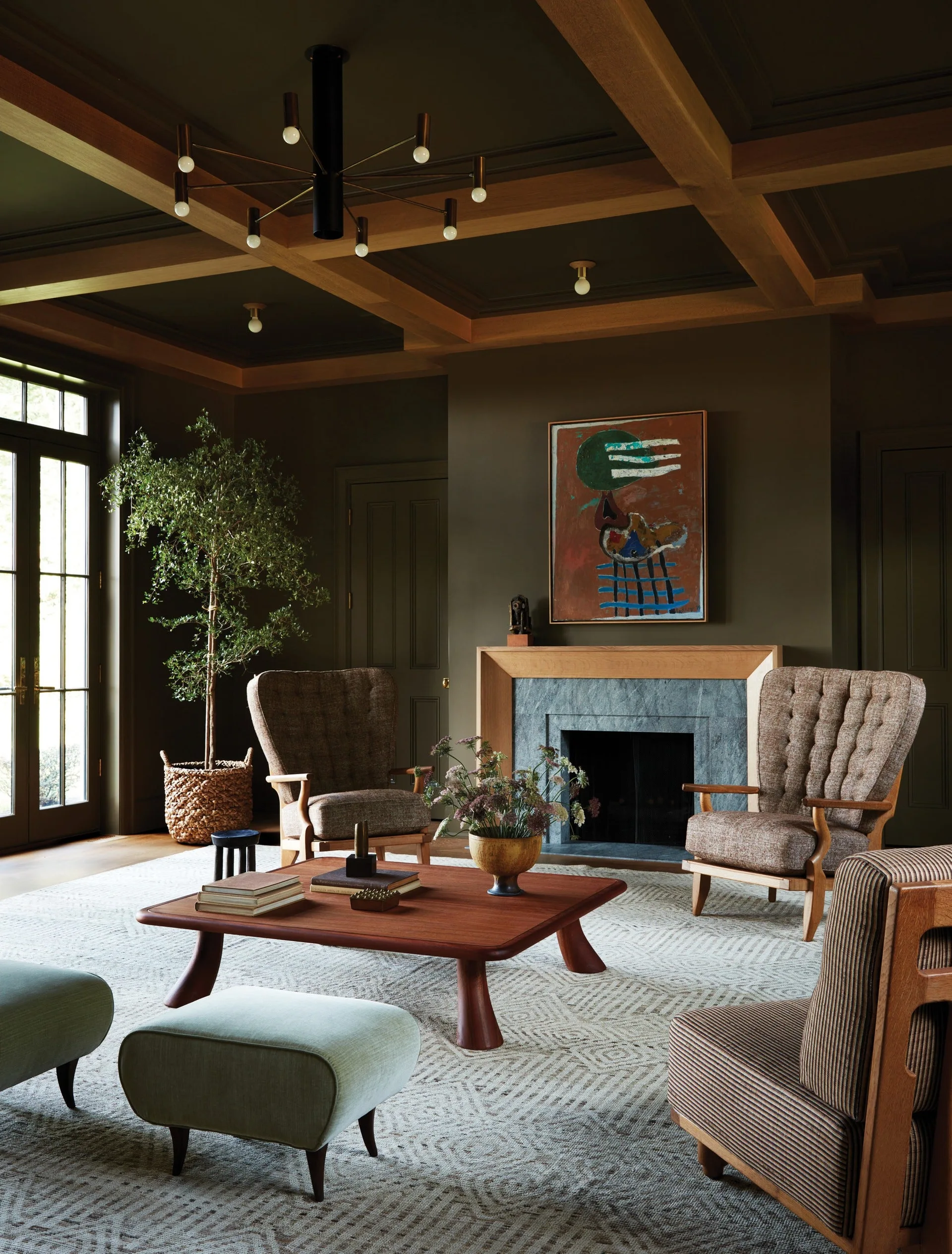 An olive green living room features a wood coffered ceiling, mid-century armchairs, a low wooden coffee table, and a stone-faced fireplace.