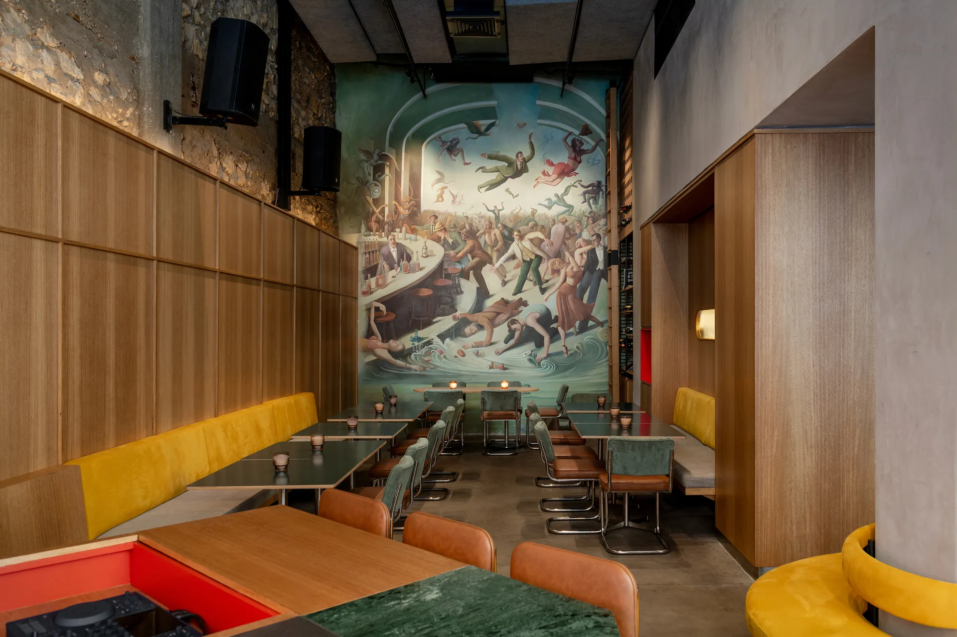 A restaurant interior features warm wood-paneled walls, long yellow booth seating, dark dining tables, and a large colorful mural on the back wall.