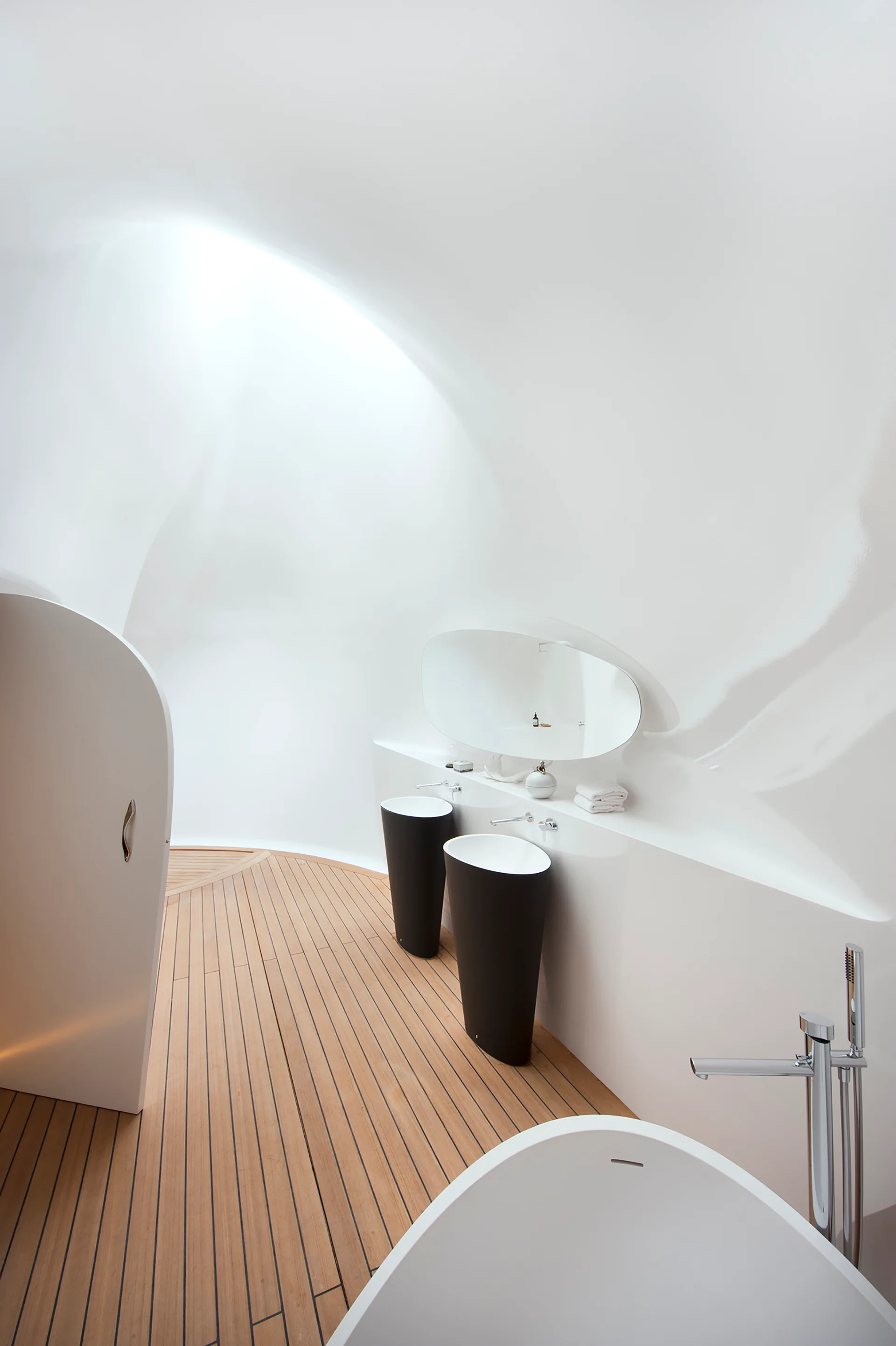 A minimalist bathroom featuring curved white walls, teak plank decking floors, two black pedestal sinks, and a large oval mirror.