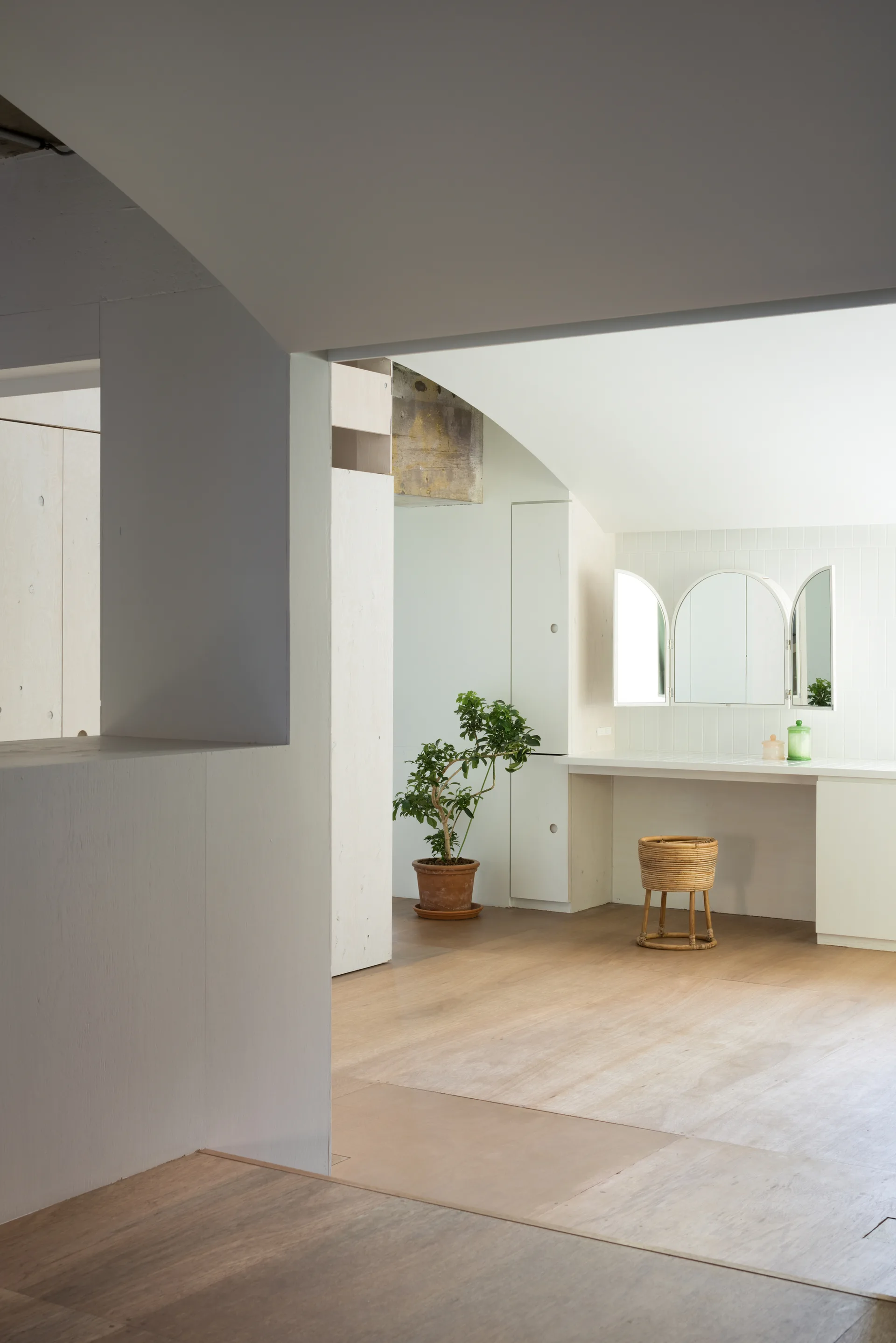 A minimalist dressing area features light wood flooring, integrated plywood cabinetry, a built-in vanity with a three-panel mirror, and a small woven stool.