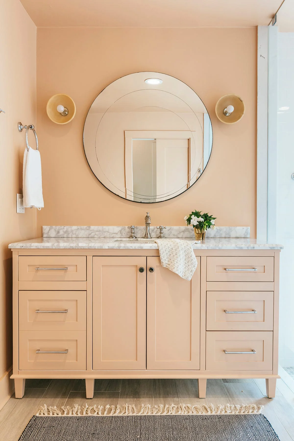 A peach-painted bathroom vanity with a white marble countertop is paired with matching peach walls, a large circular mirror, and two round sconces.