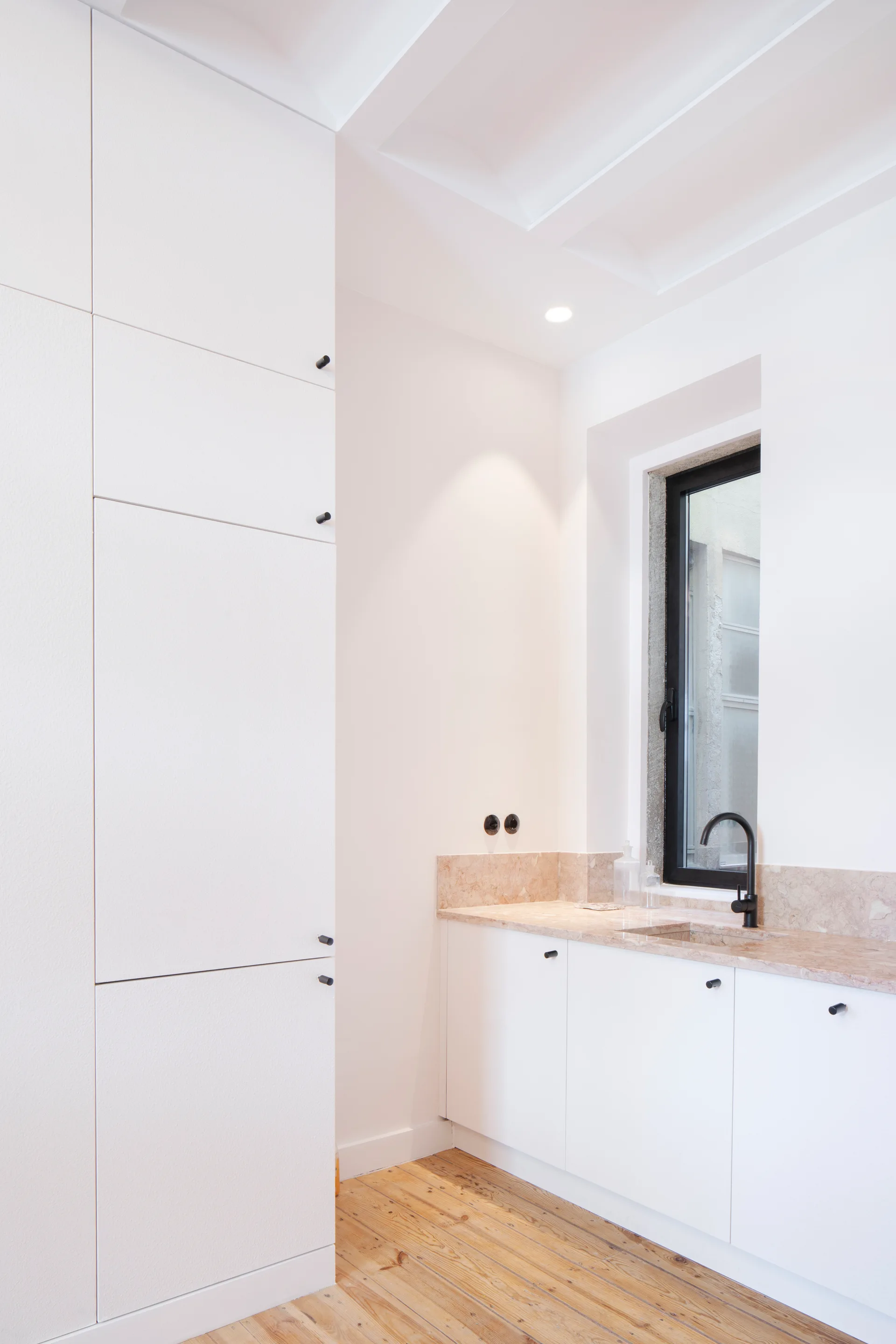 A bathroom features tall white cabinets, a beige stone countertop with an integrated sink, a black faucet, and natural wood flooring.