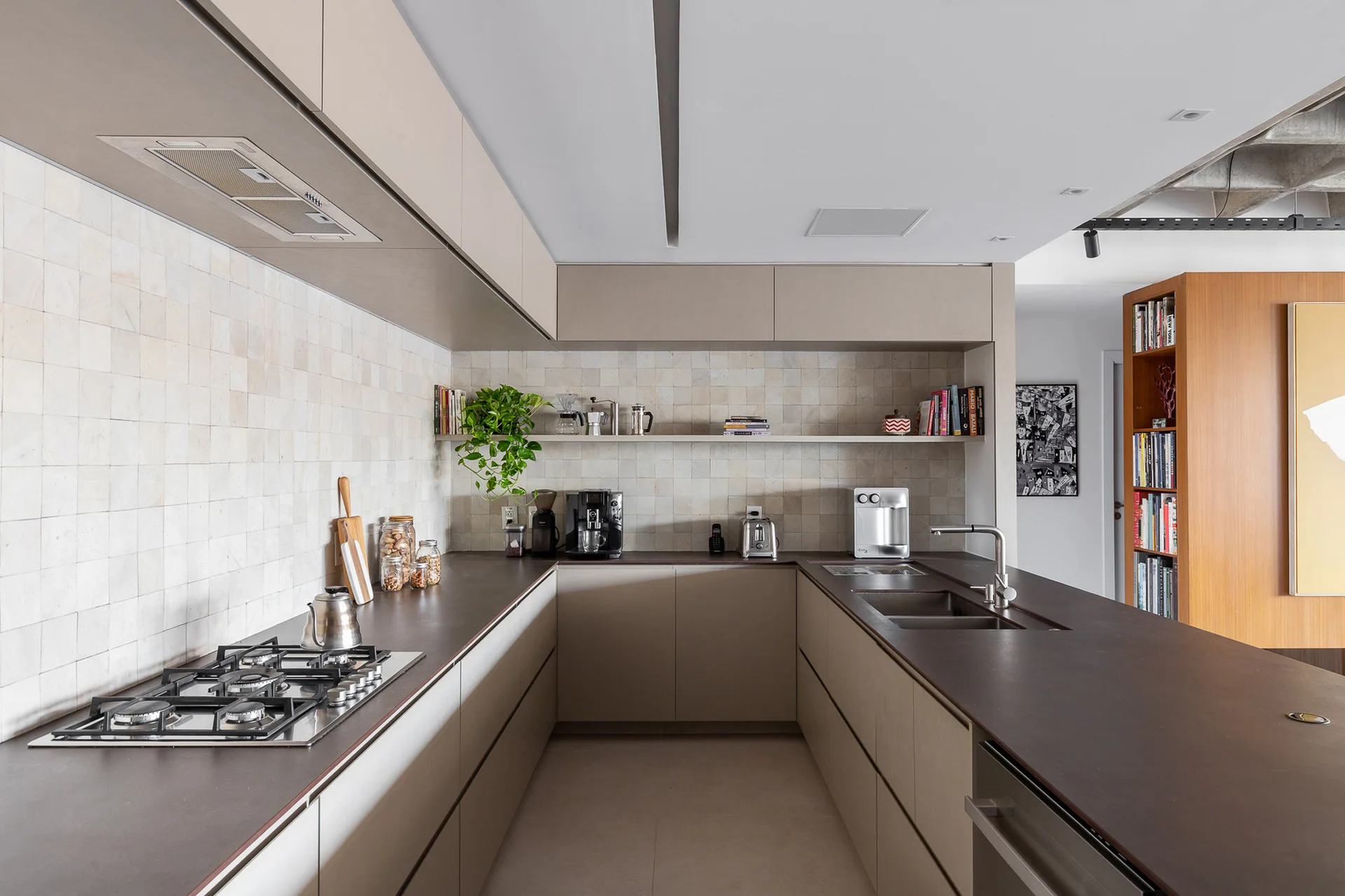 A modern U-shaped kitchen with matte beige cabinets, dark brown countertops, and a light square-tiled backsplash featuring a single floating shelf.