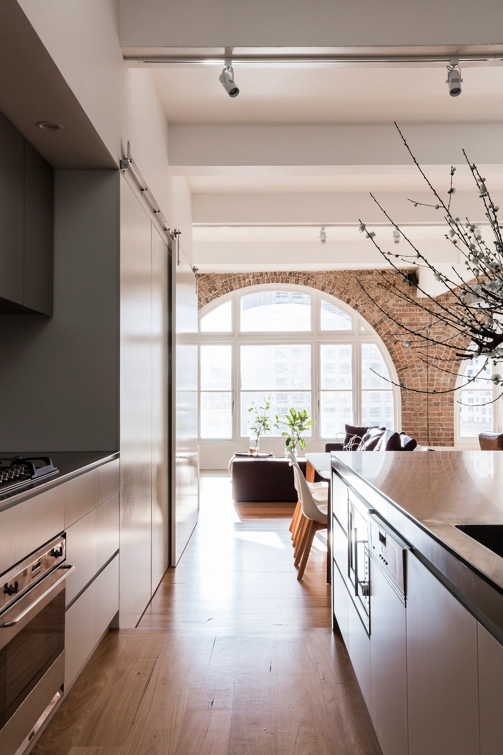 A modern kitchen with a gray island leads to an open dining and living area framed by a large arched window with exposed brick.