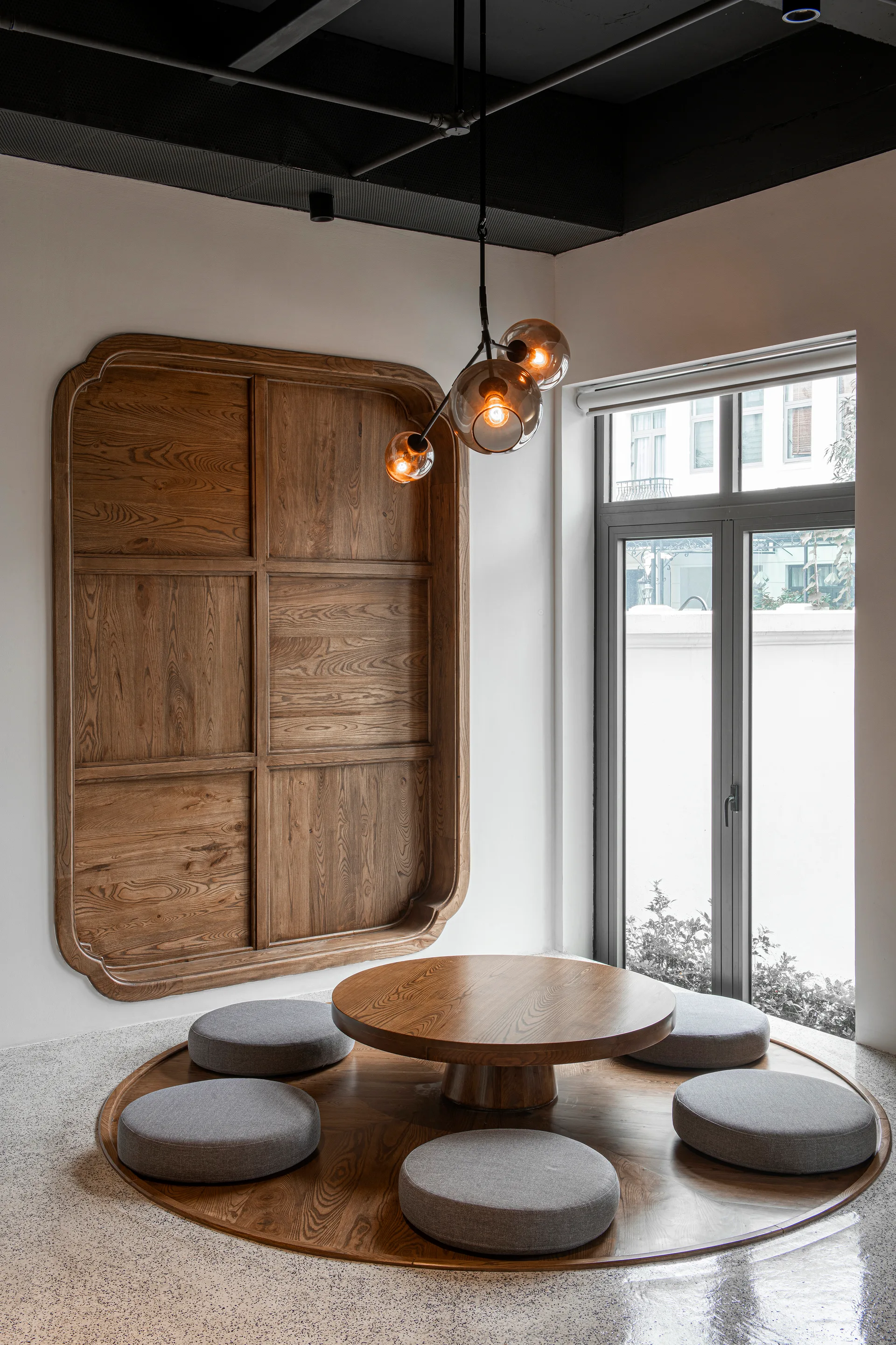 A low wooden round table with grey floor cushions sits on a circular wooden platform next to a matching wall panel and a glass pendant light.