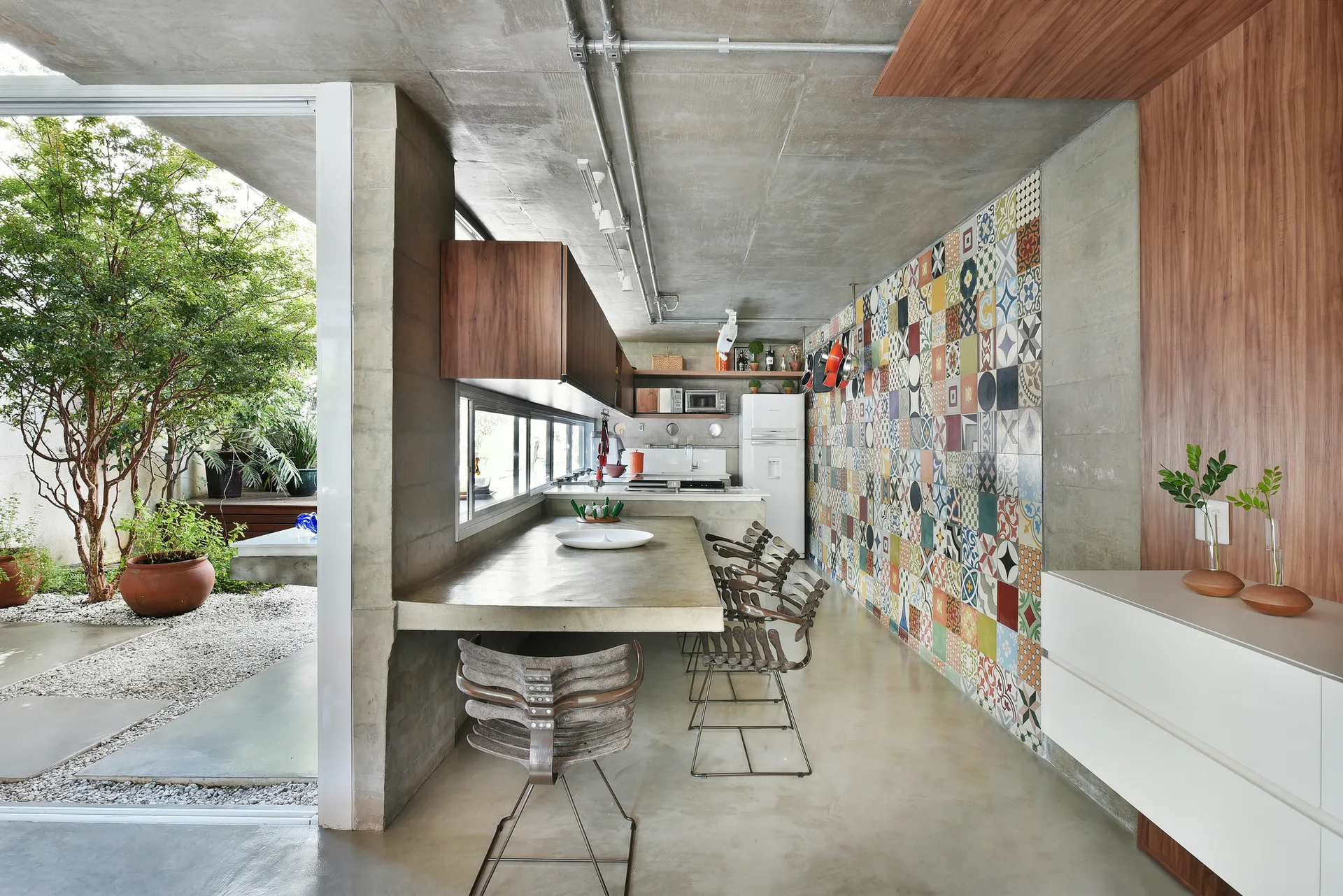 An industrial kitchen features a concrete dining counter, wood cabinets, a colorful patchwork tile wall, and glass doors opening to a garden courtyard.