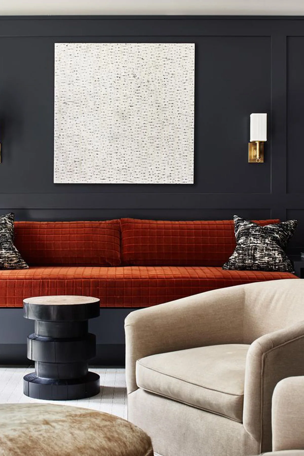 A living room features a burnt orange tufted sofa set against a dark charcoal paneled wall decorated with textured square art and a brass sconce.