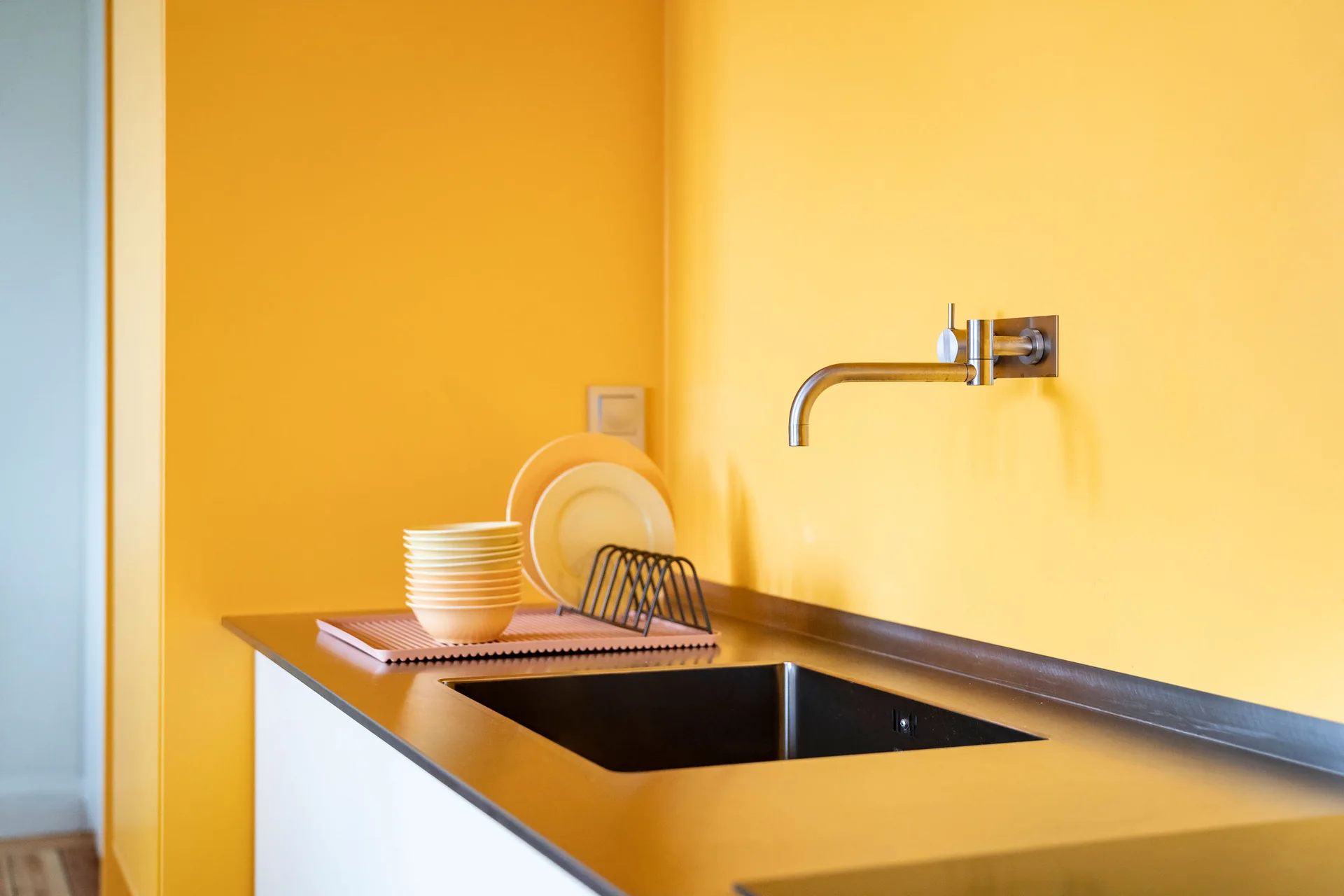 A wall-mounted metal faucet sits above an inset black sink on a stainless steel countertop against a vibrant yellow wall with drying dishes.