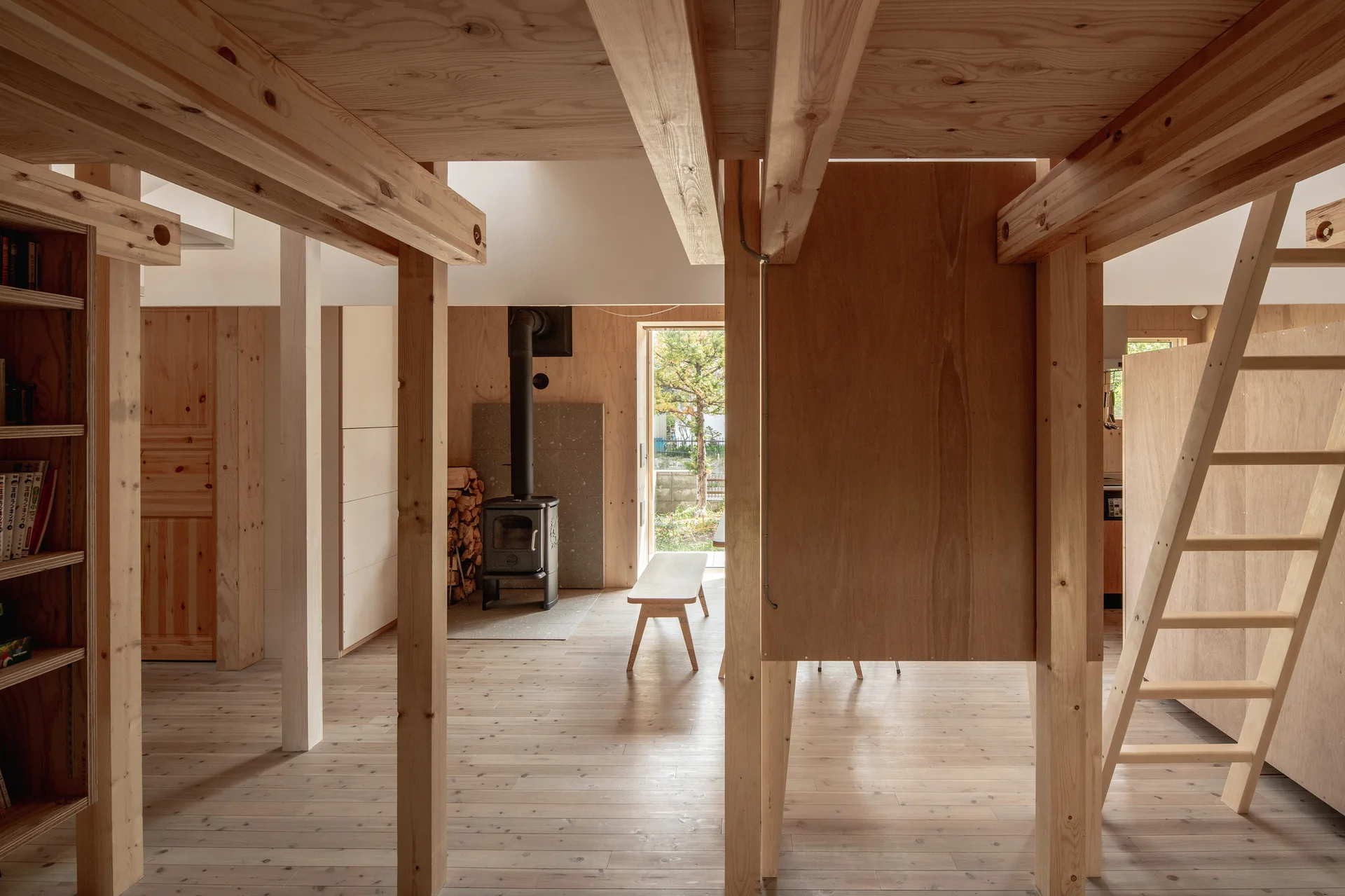 A minimalist cabin interior with light plywood walls, exposed structural timber beams, a wooden ladder, and a black wood-burning stove.