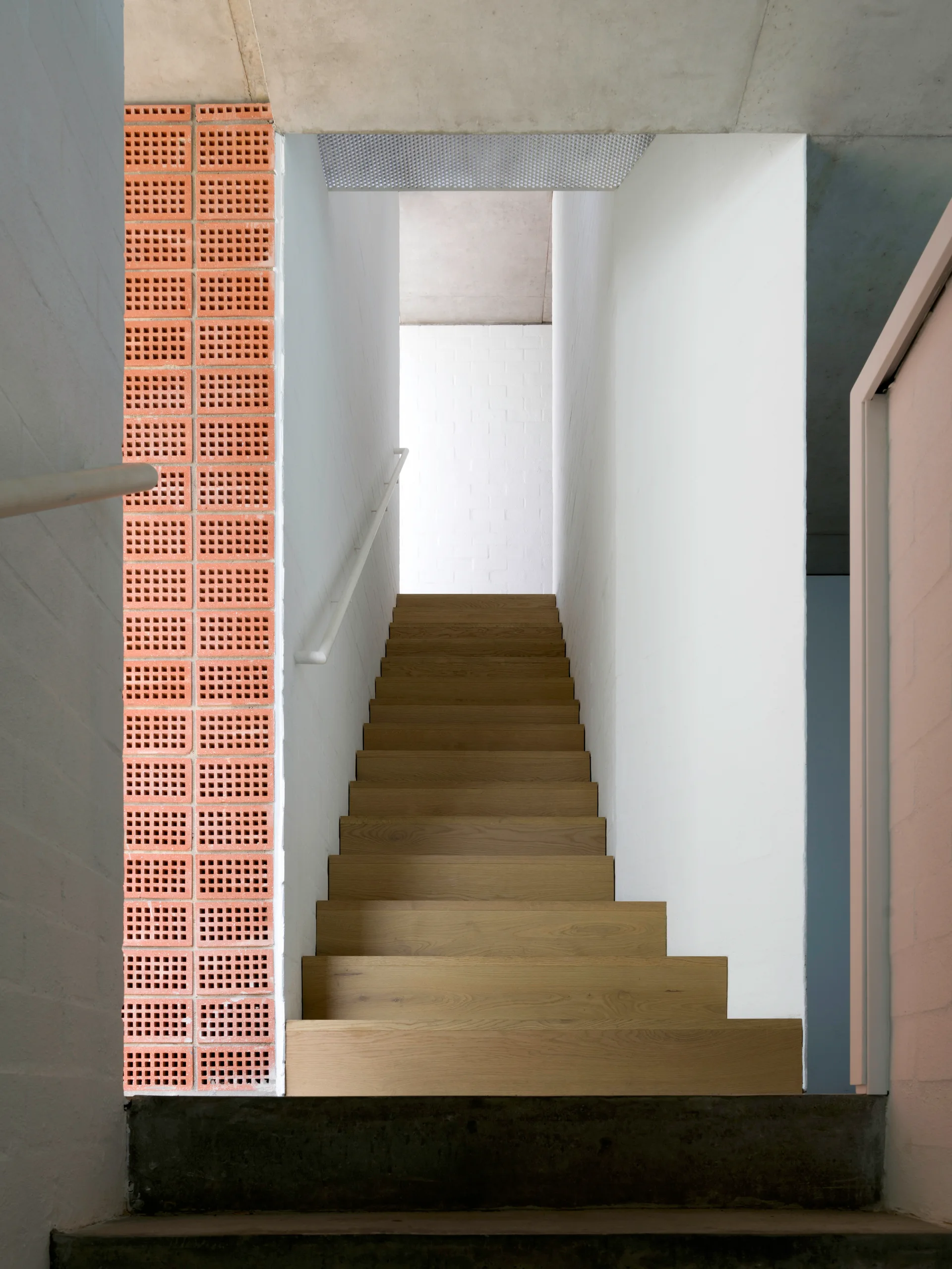 A straight wooden staircase with light oak treads rises between smooth white walls and a vertical column of red perforated terracotta bricks.