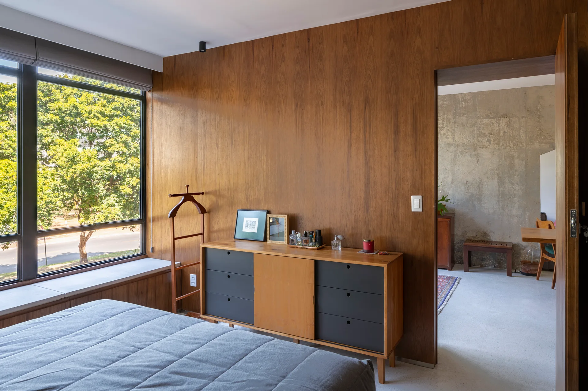 A modern bedroom featuring vertical wood wall paneling, a mid-century wooden credenza, and an open doorway looking into a concrete room.