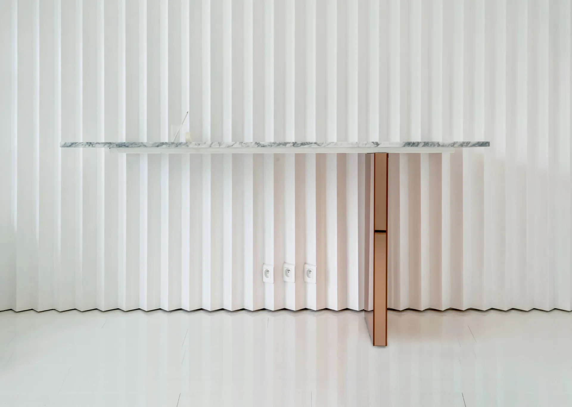 A minimalist white marble console shelf with a single polished copper leg is mounted to a white fluted wall.