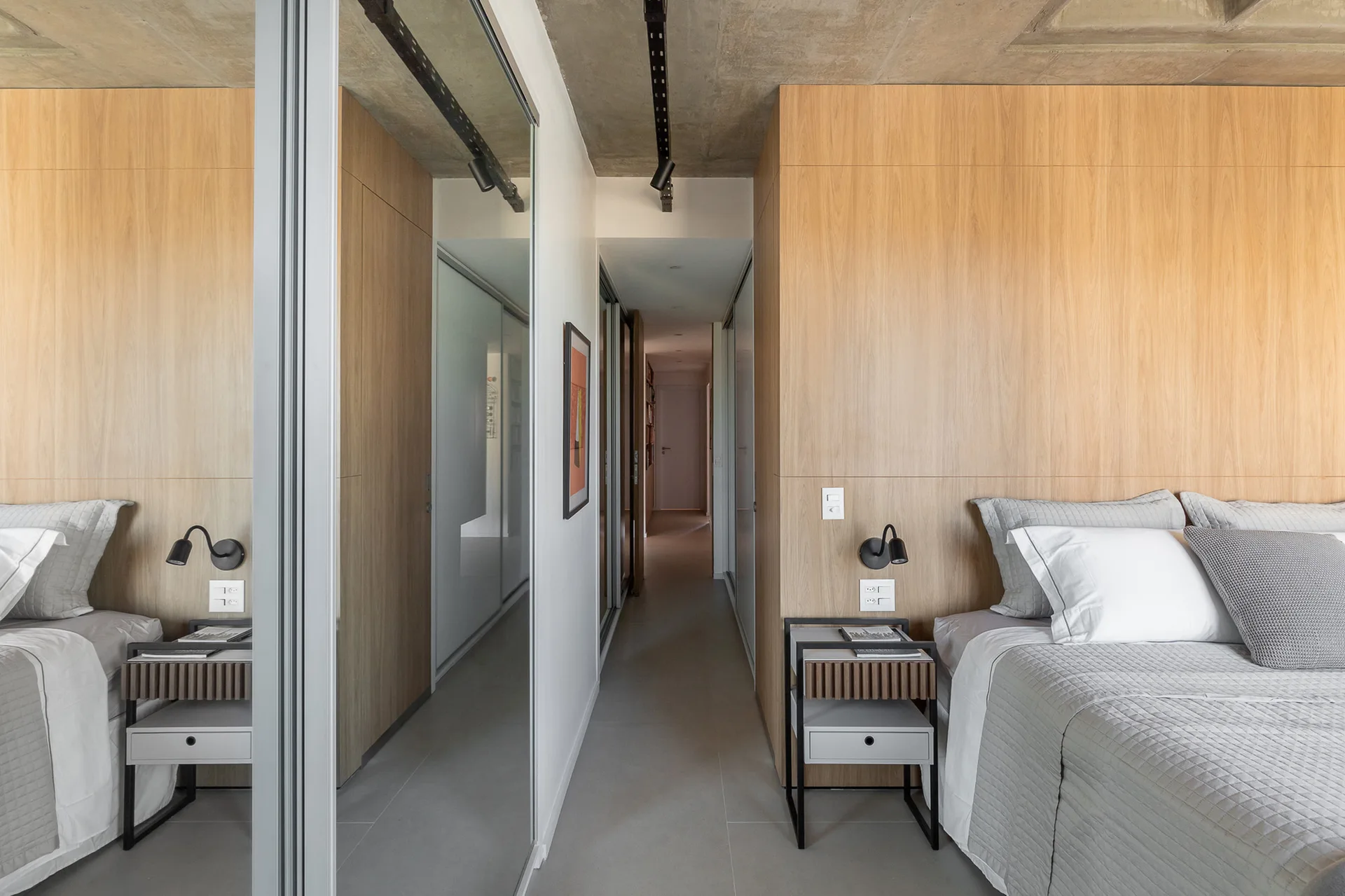 A minimalist bedroom featuring a warm wood paneled wall behind the bed, an exposed concrete ceiling, a grey bedspread, and a mirrored closet wall.