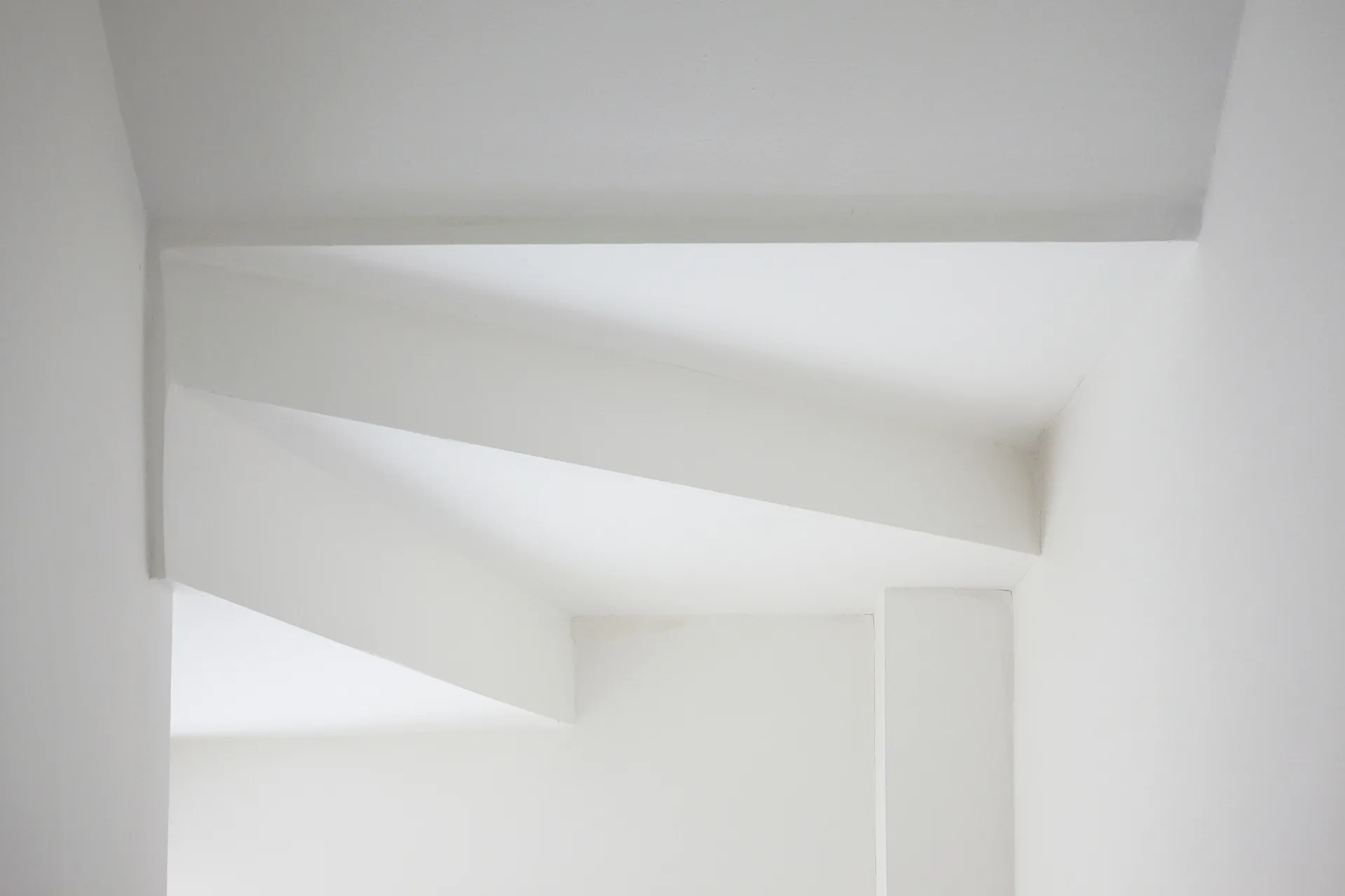 A close-up of a minimalist white interior corner featuring smooth matte walls and a sharp architectural ceiling beam.