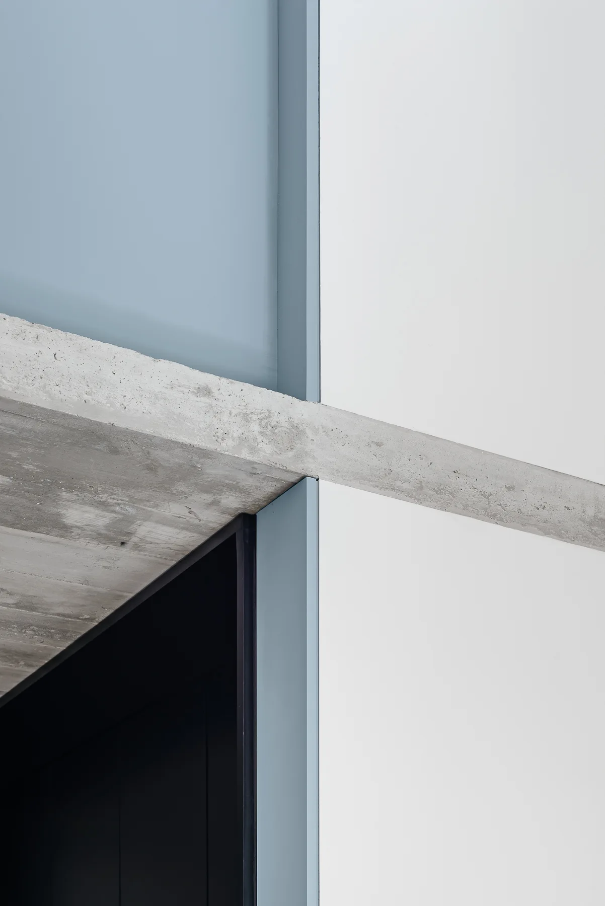 A close-up of a minimalist architectural junction featuring an exposed grey concrete beam crossing a muted blue vertical frame against white walls.