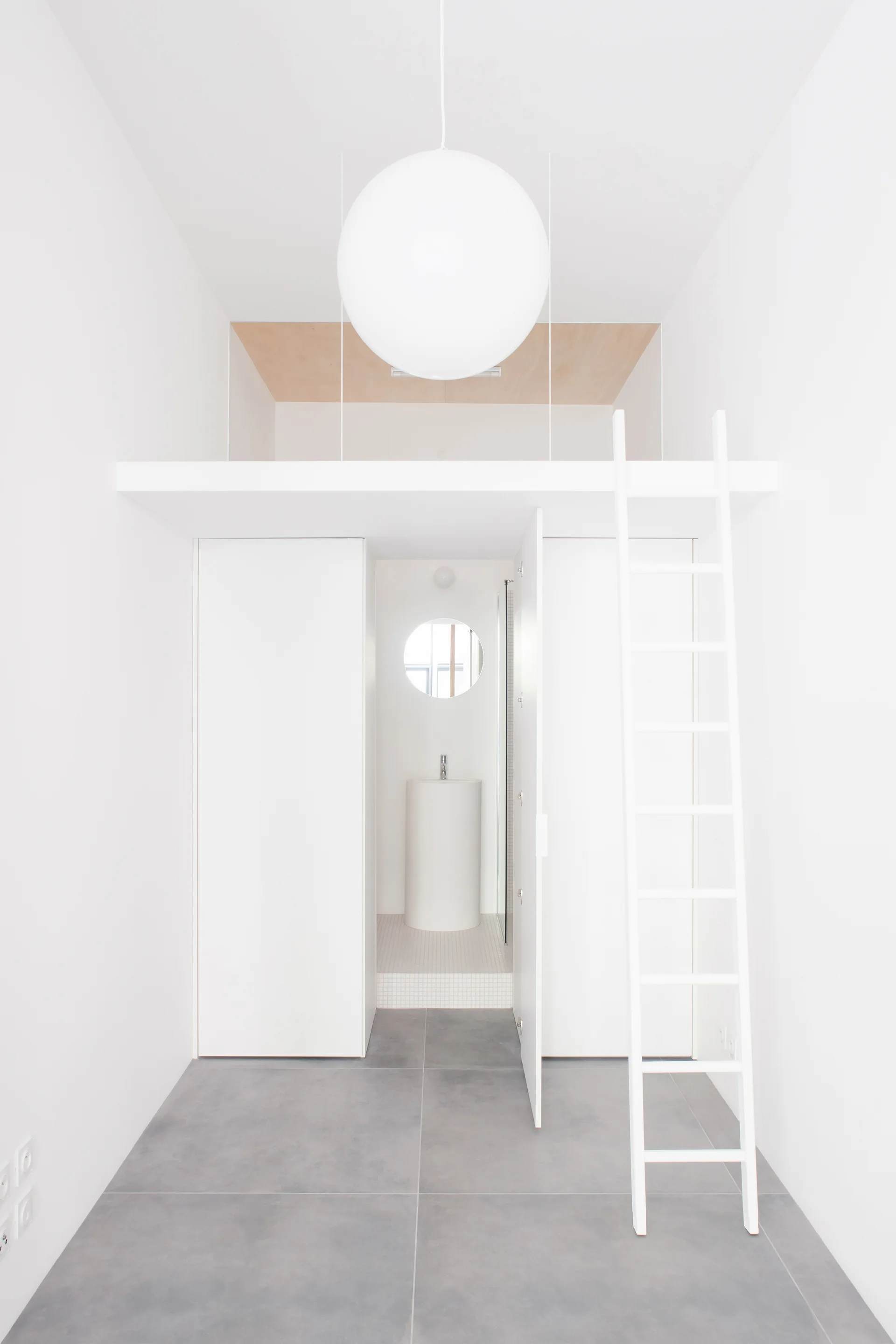 A minimalist white interior with a gray tile floor, featuring a ladder leading to a loft and an open door to a small bathroom alcove.