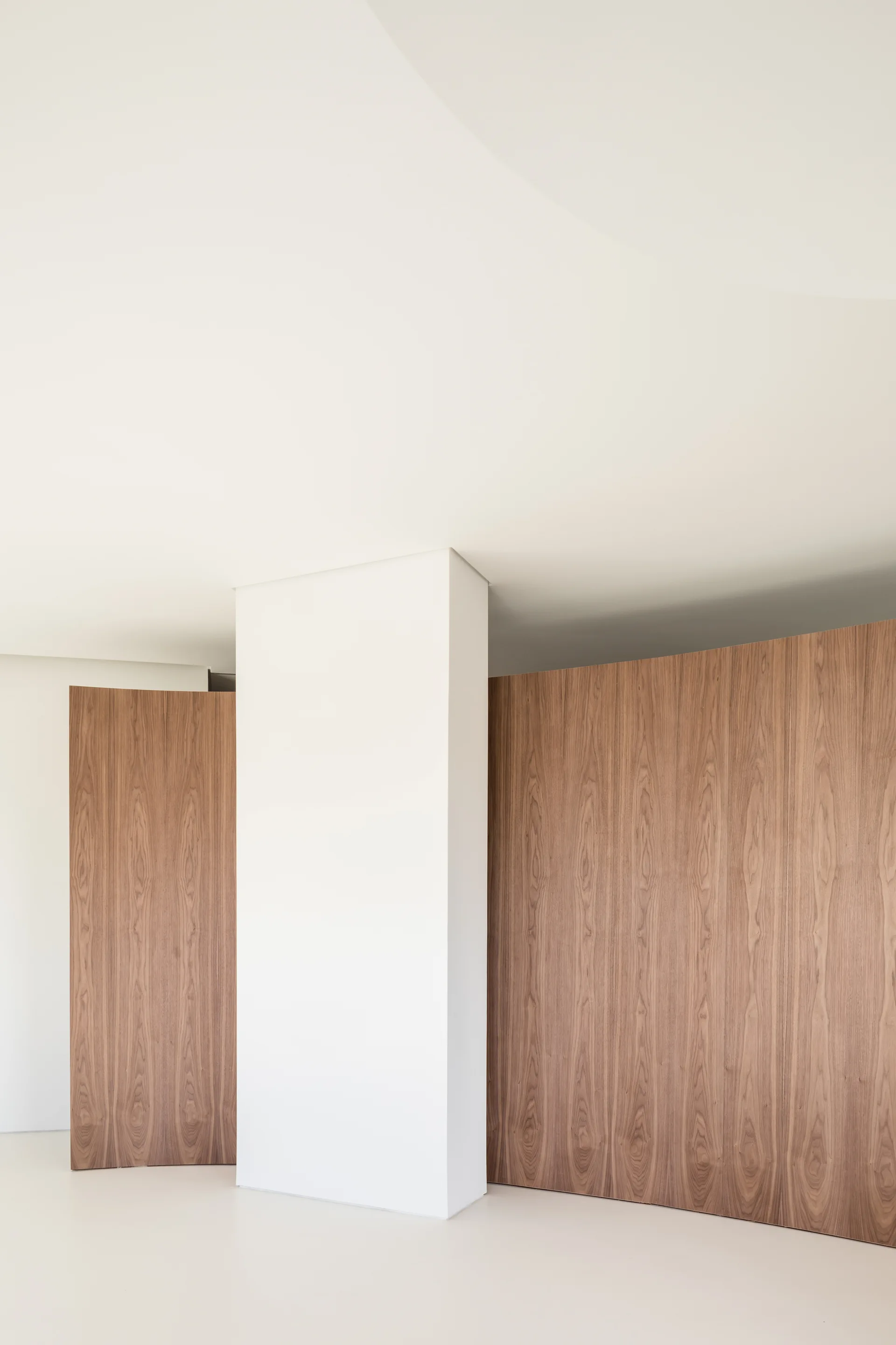 Minimalist white architectural column flanked by curved wood paneling with a natural vertical grain against a clean white ceiling and floor.