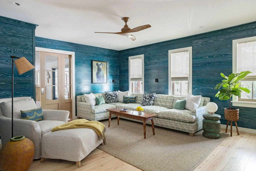 A living room featuring blue wood-grain wallpaper, a light-colored patterned sectional sofa, a wooden coffee table, and a woven rug.