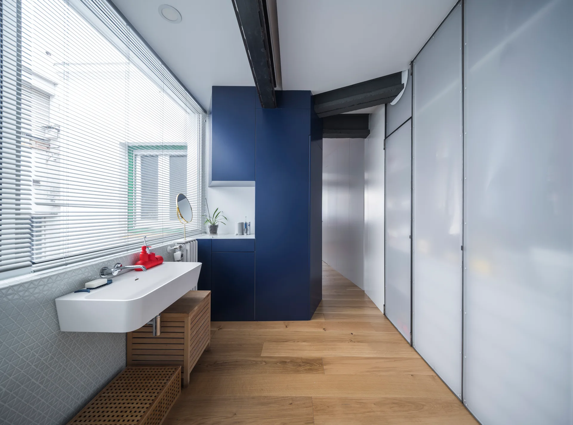 A modern bathroom with a white wall-mounted sink, a slatted wood vanity, deep blue storage cabinets, and frosted glass sliding doors.