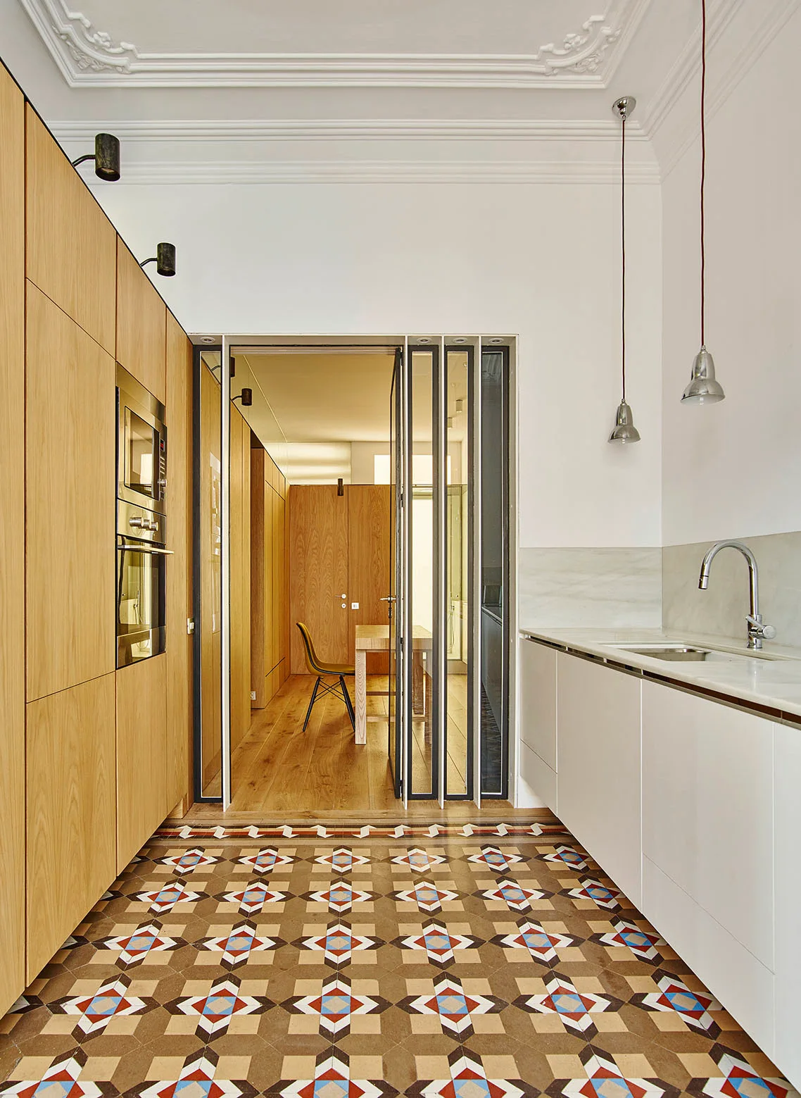A modern kitchen with tall light wood cabinets, white lower cabinets, and a colorful geometric tile floor underneath an ornate plaster ceiling.