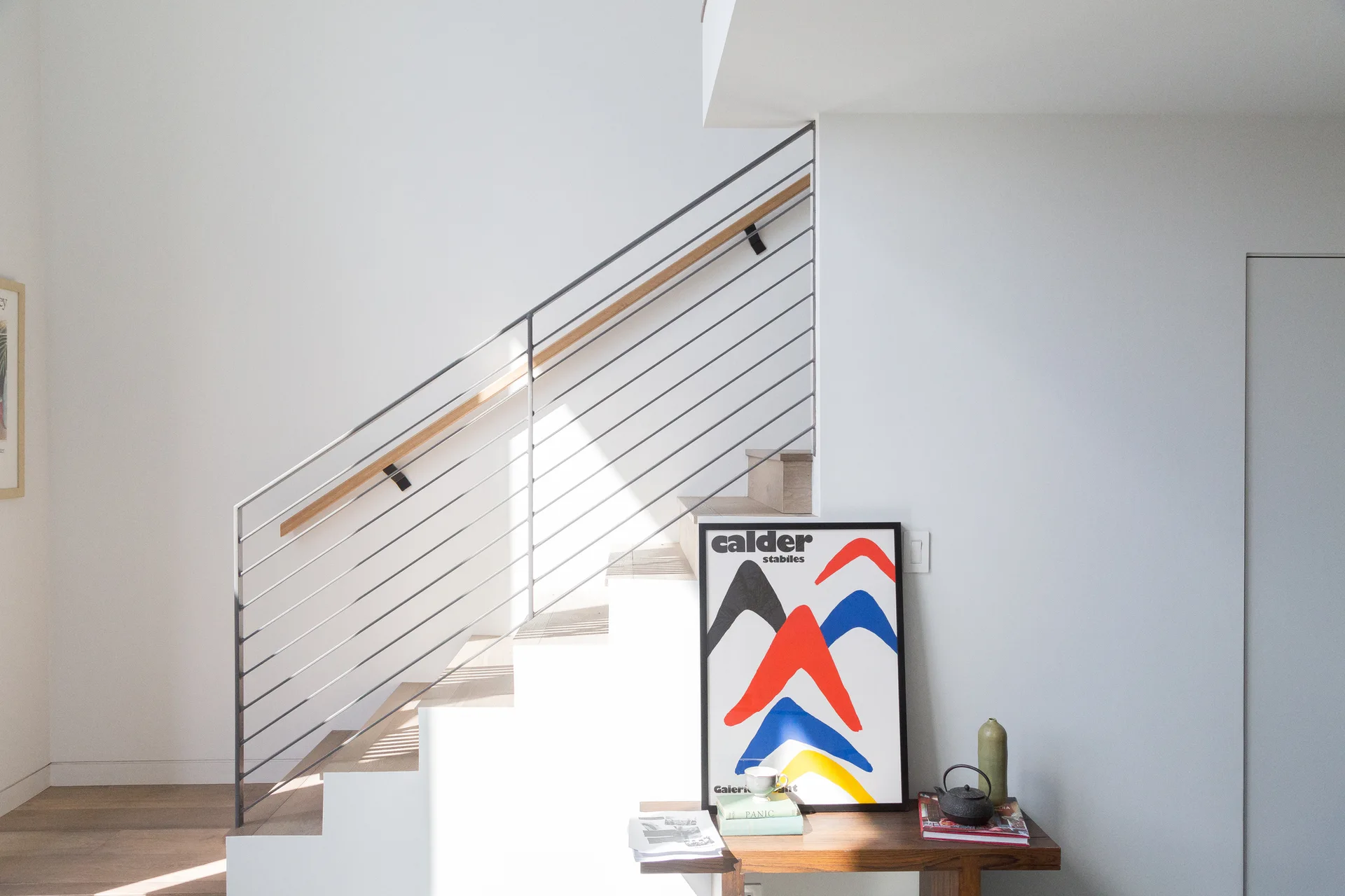 A minimalist staircase features light wood treads and a thin steel rod railing, with a wooden console table displaying art and decor alongside it.