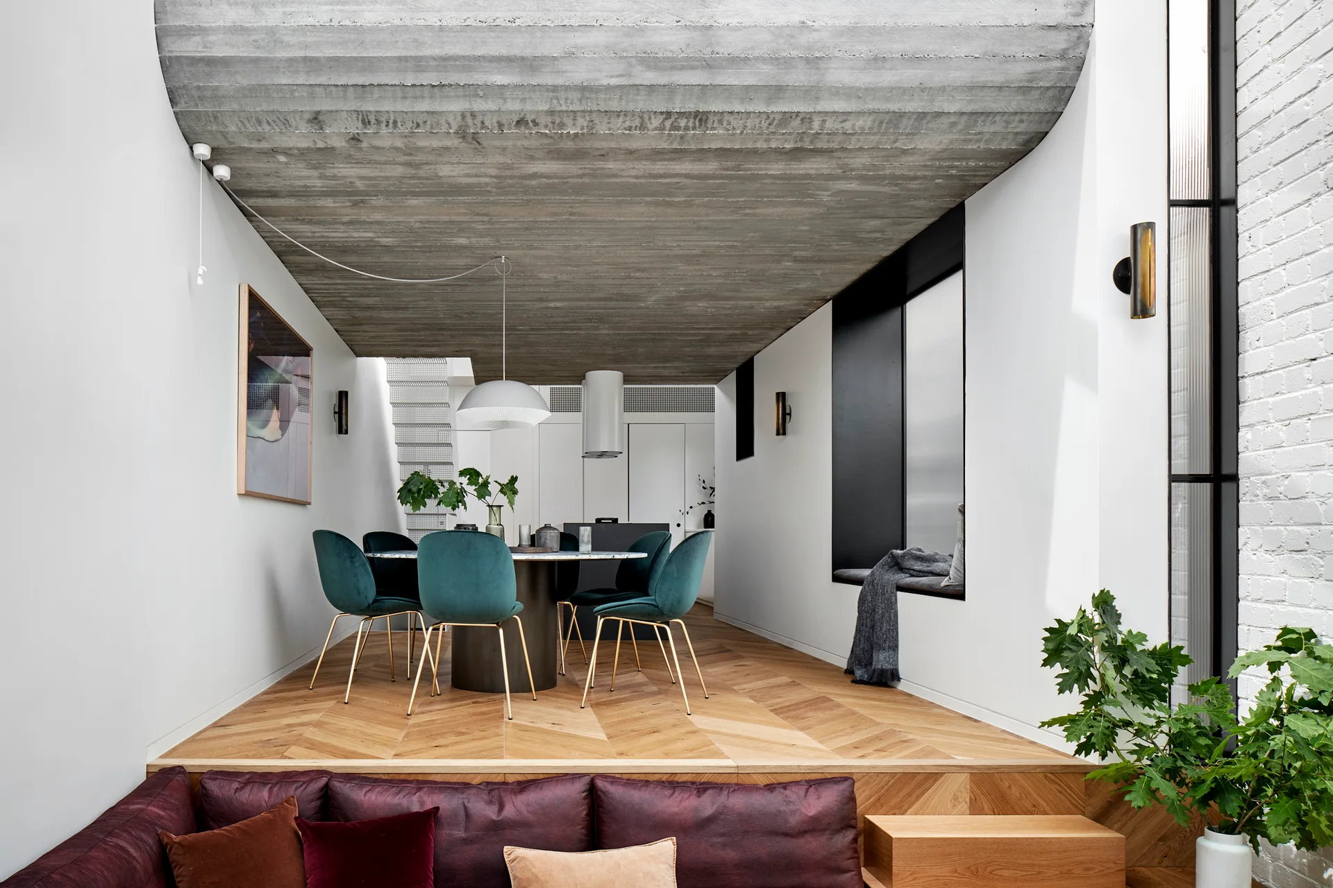 A split-level room features a raised dining area with teal velvet chairs and a chevron floor under an exposed concrete ceiling.