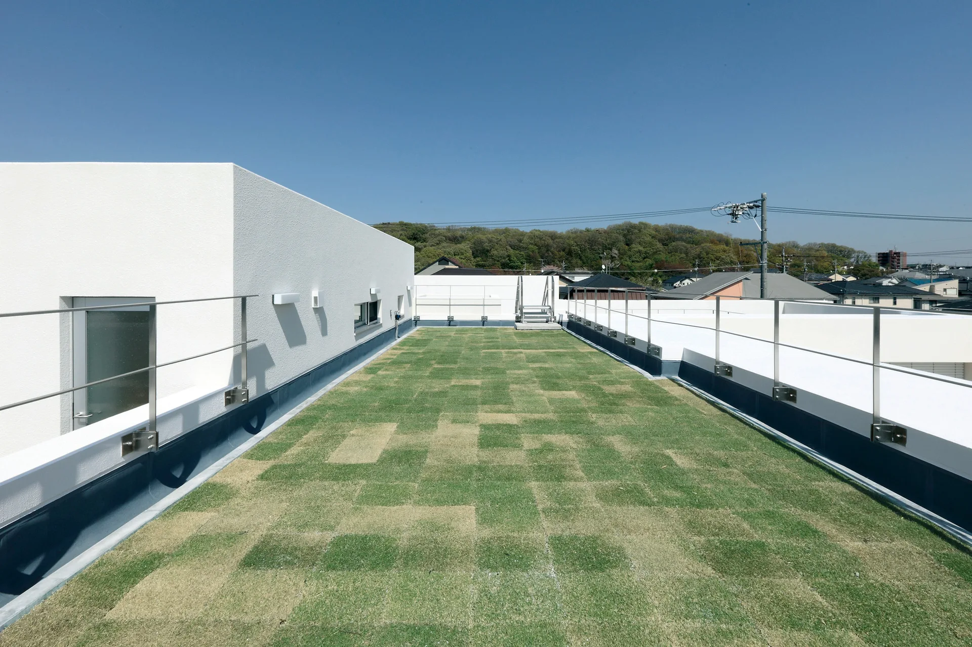 A long rooftop terrace features checkered green grass tiles, white stucco walls, and metal railings under a clear blue sky.
