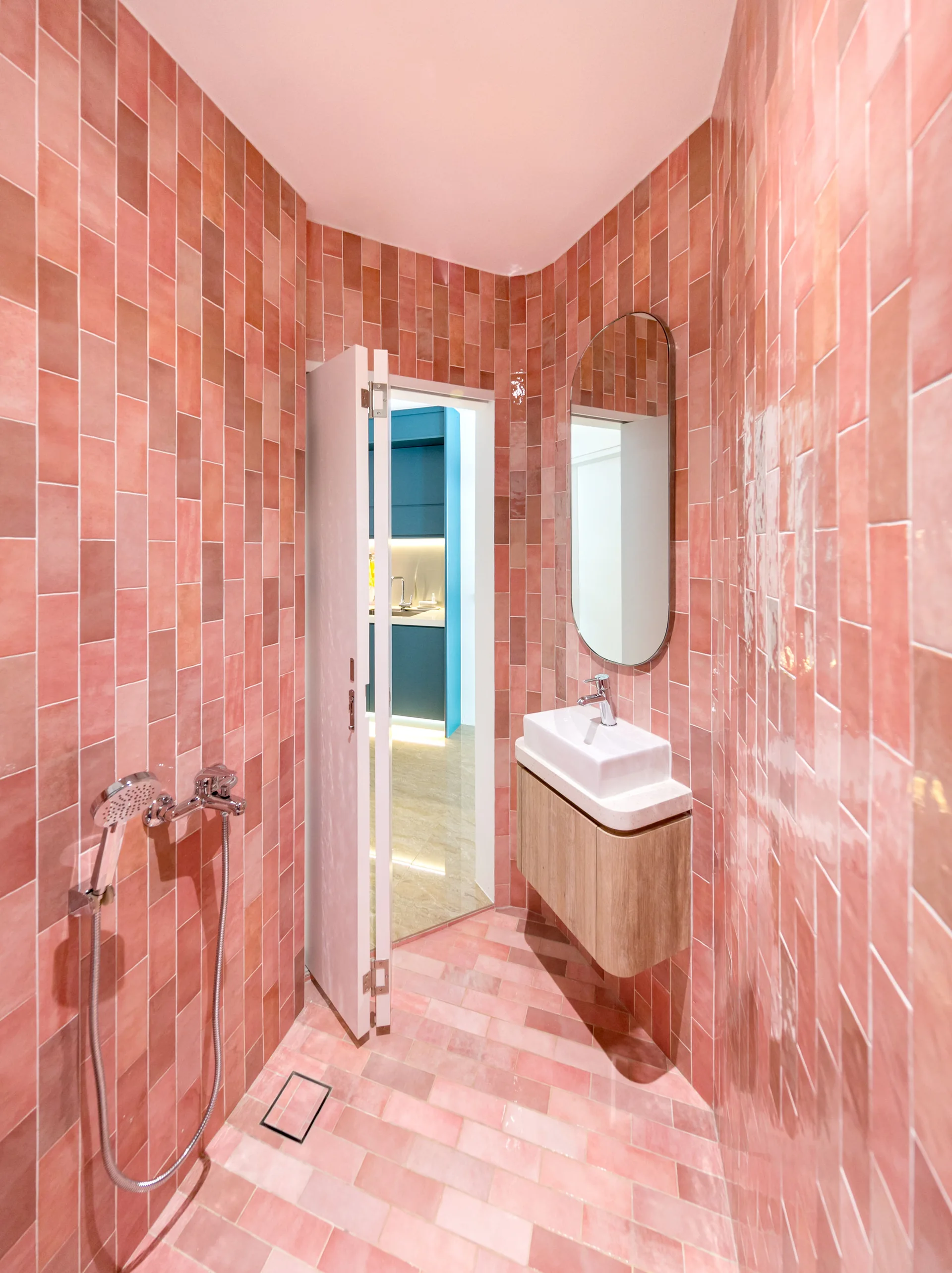 A modern bathroom features glossy pink vertical tiles on the walls, matching floor tiles, a floating wooden vanity, and an oval mirror.
