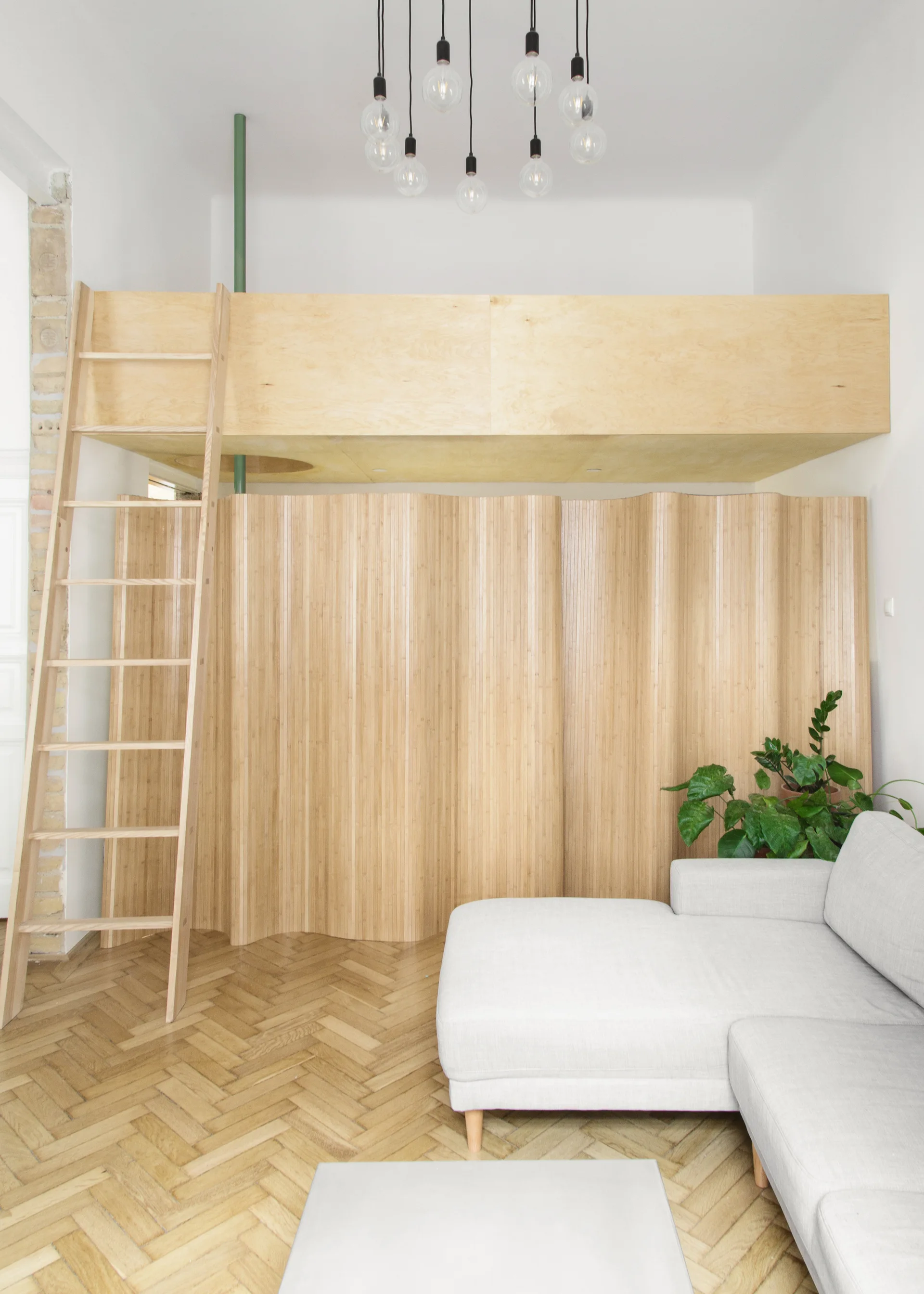 A light grey sectional sofa sits on a herringbone wood floor next to a wavy fluted wood partition beneath a light plywood loft with a ladder.
