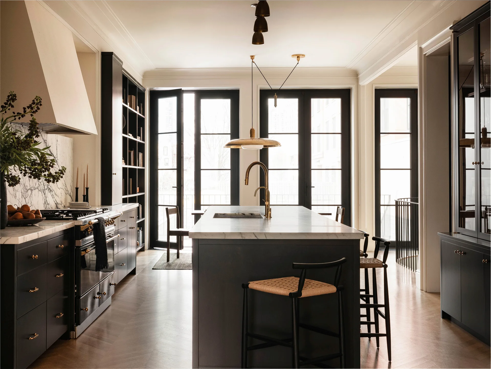 A transitional kitchen features black cabinetry, a white marble island with woven seat stools, and tall black-framed French doors.