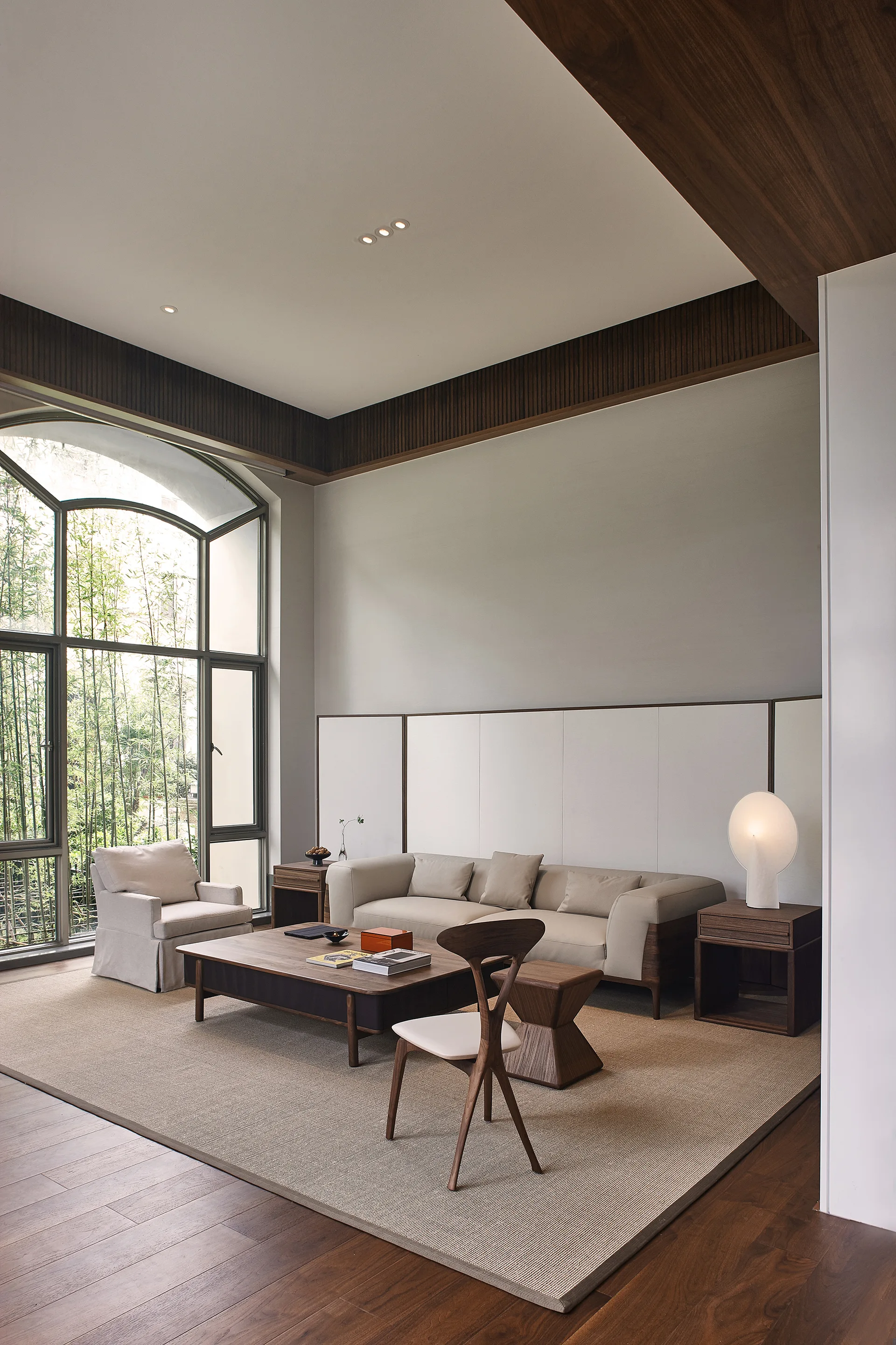 A contemporary living room featuring a beige sofa, low wood tables, a curved accent chair, and a large arched window looking out at green bamboo.
