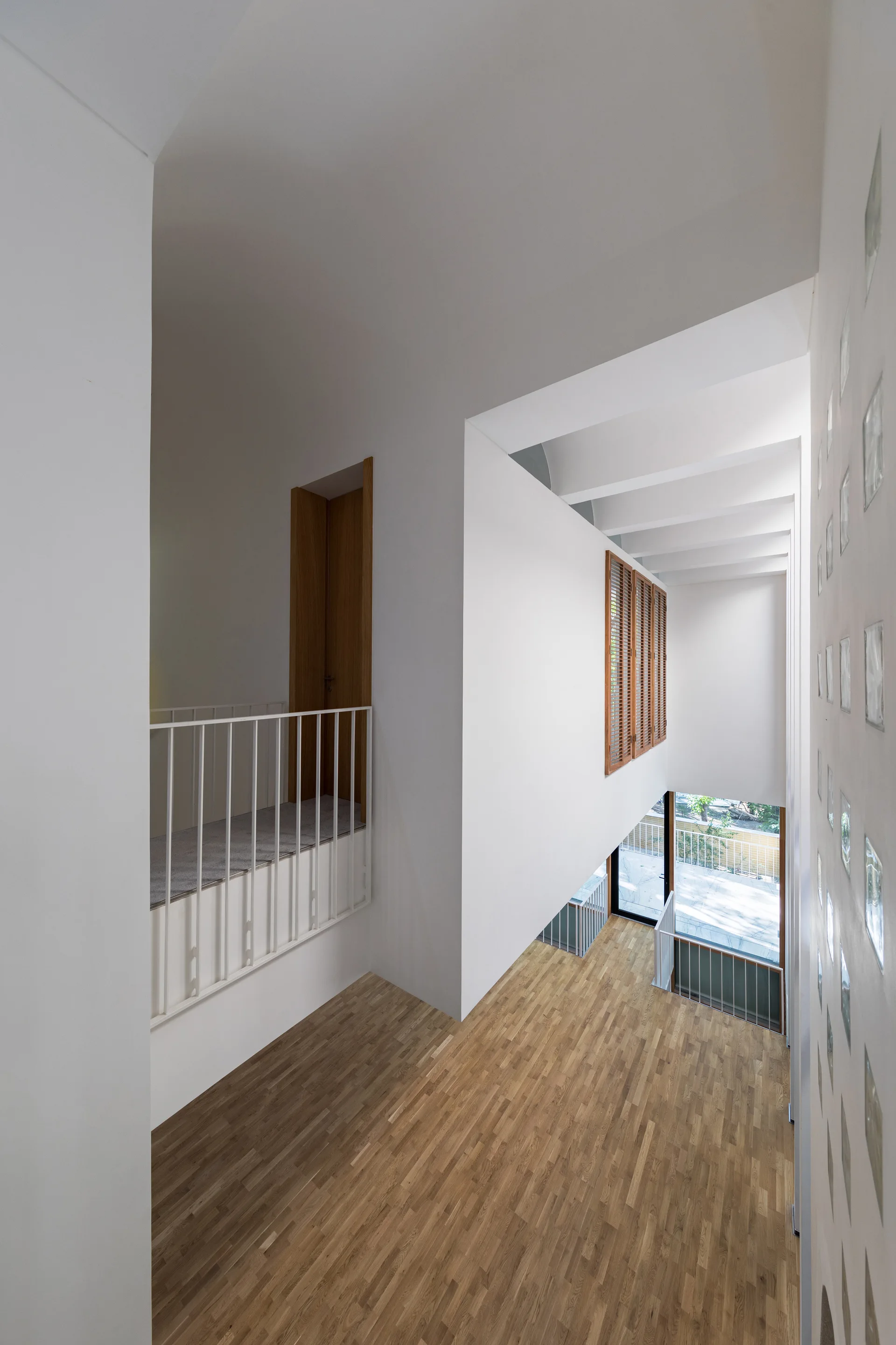 A multi-level interior hallway features light wood flooring, white walls, metal railings, a wooden shuttered window, and a glass block wall.