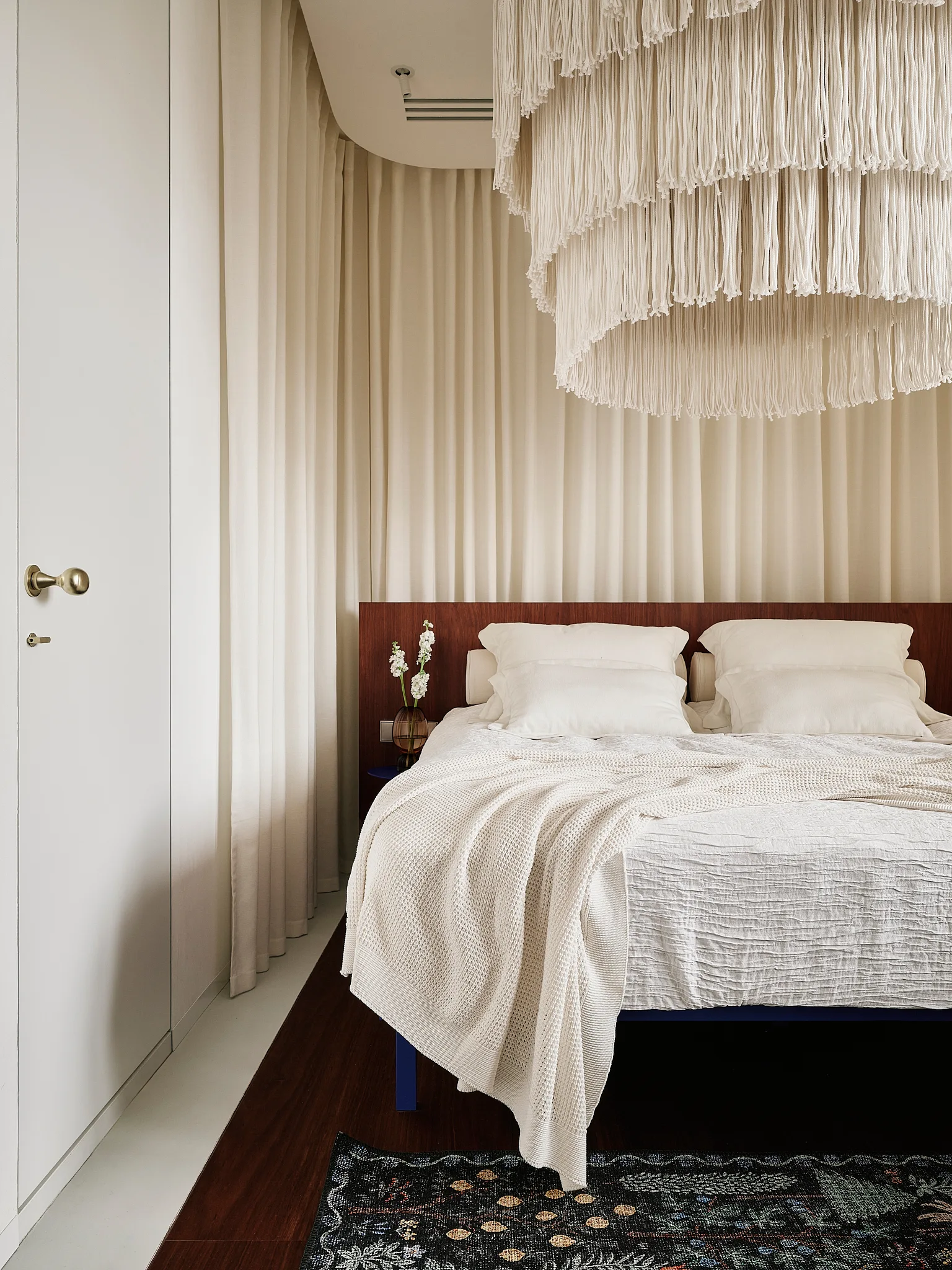A minimalist bedroom featuring a bed with white bedding, a dark wood headboard, a large tiered fringe chandelier, and draped cream curtains.