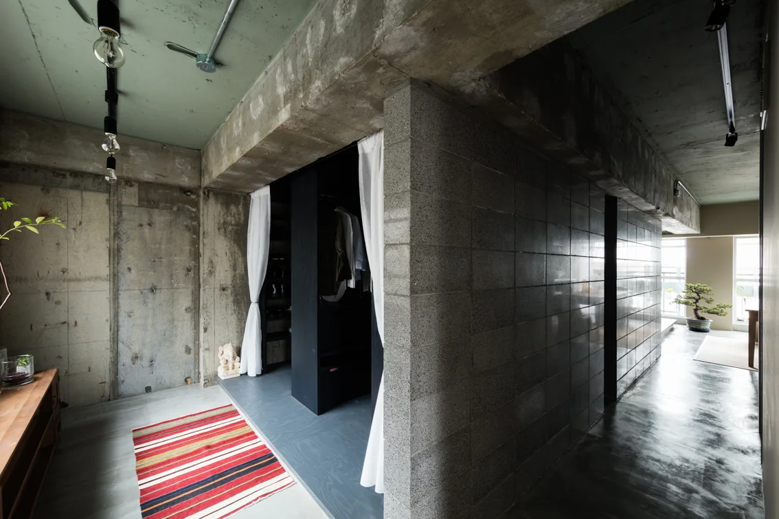 An industrial dressing room features raw concrete walls, a dark closet unit framed by white curtains, and exposed track lighting on a green ceiling.