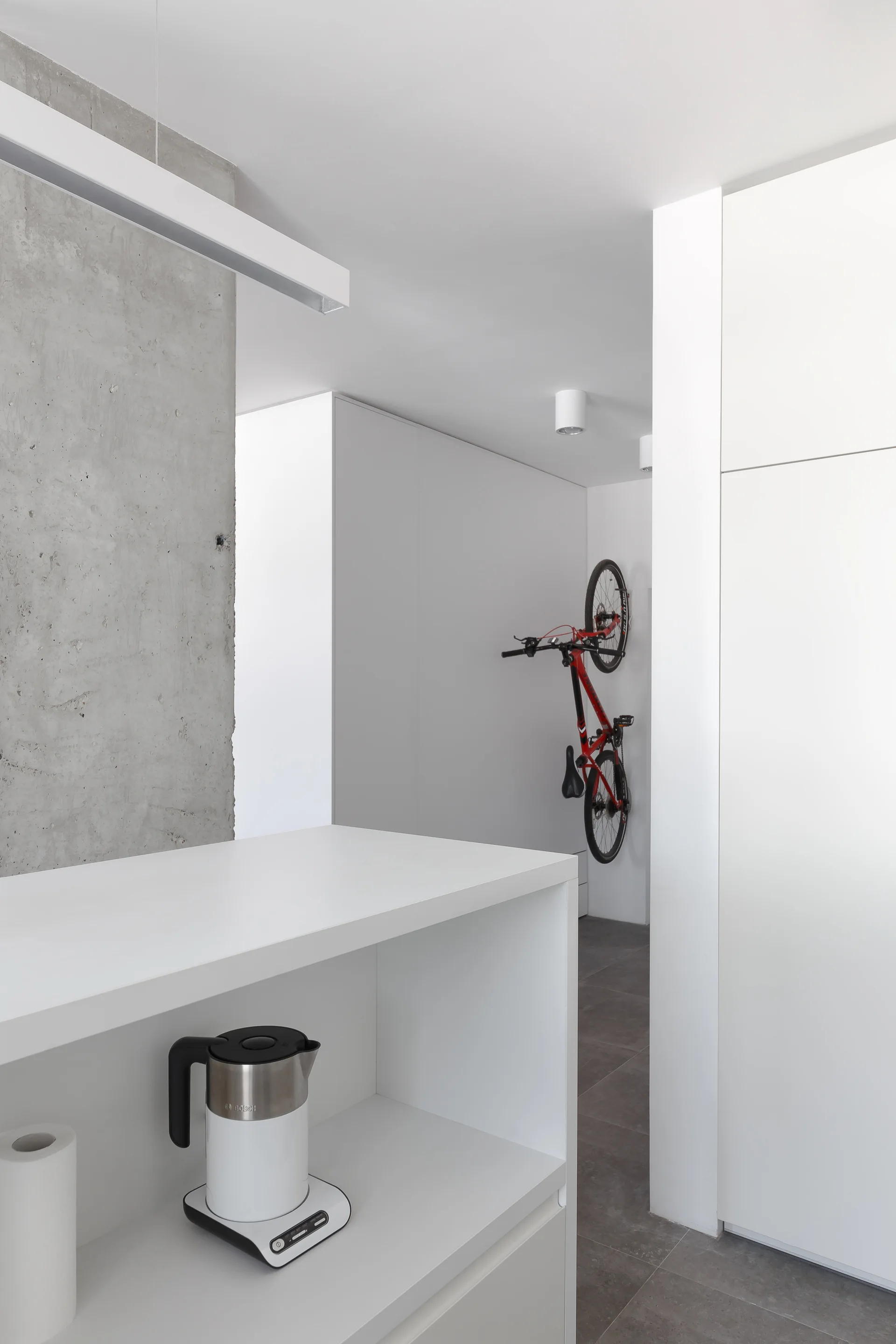 A minimalist hallway features sleek white cabinetry, an exposed grey concrete wall, and a red bicycle mounted vertically on the wall.
